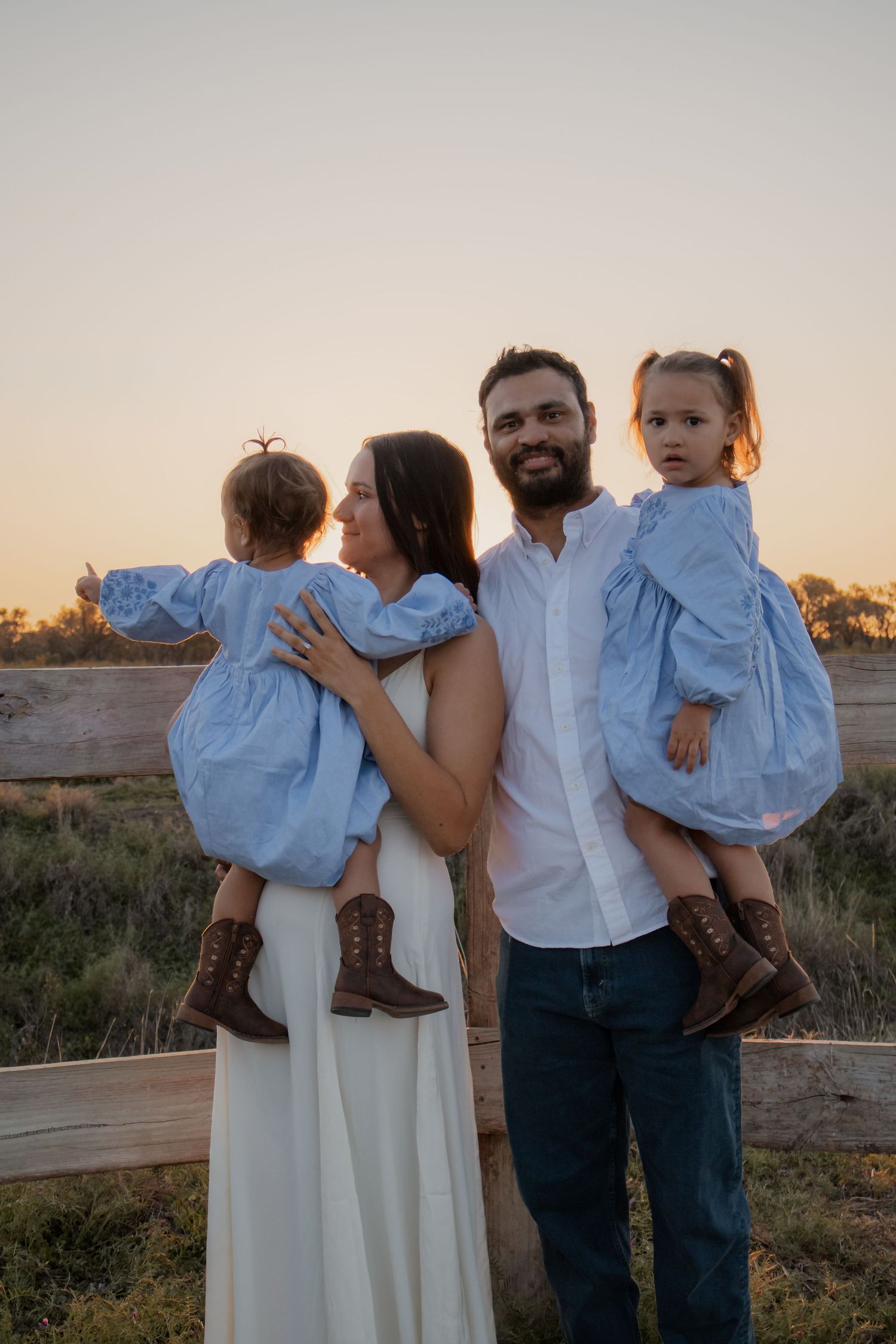 Family of four at sunset, parents holding two young girls in matching blue dresses near a wooden fence.