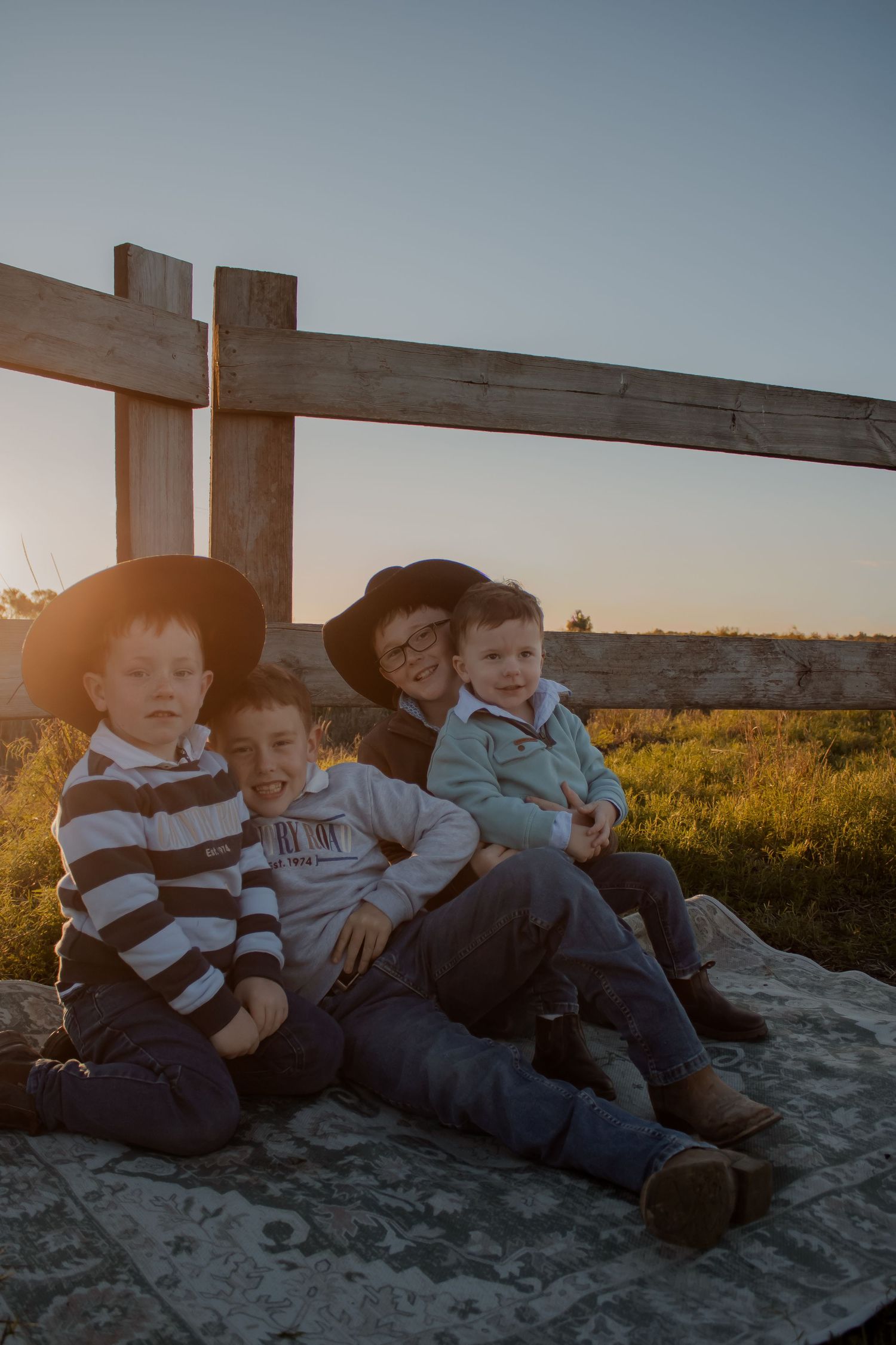 Four children sitting on a blanket by a wooden fence at sunset in a rural field, two wearing black cowboy hats.
