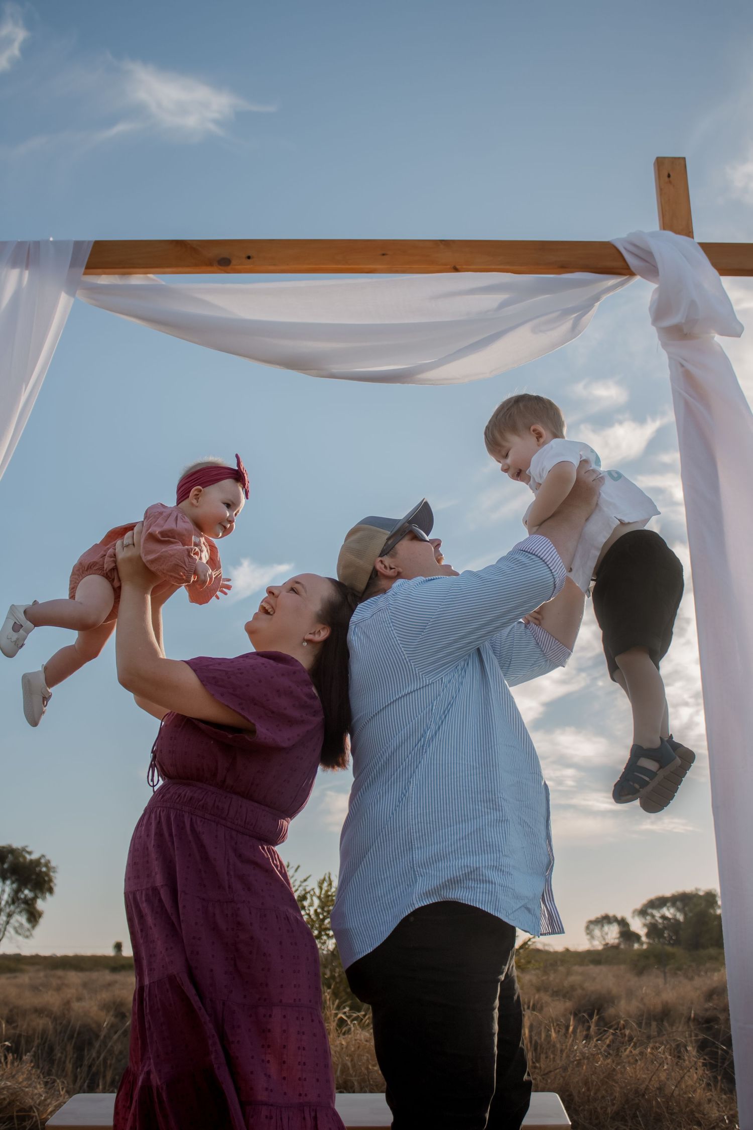 Family with two babies under a white draped wooden arch in an outdoor field at sunset.