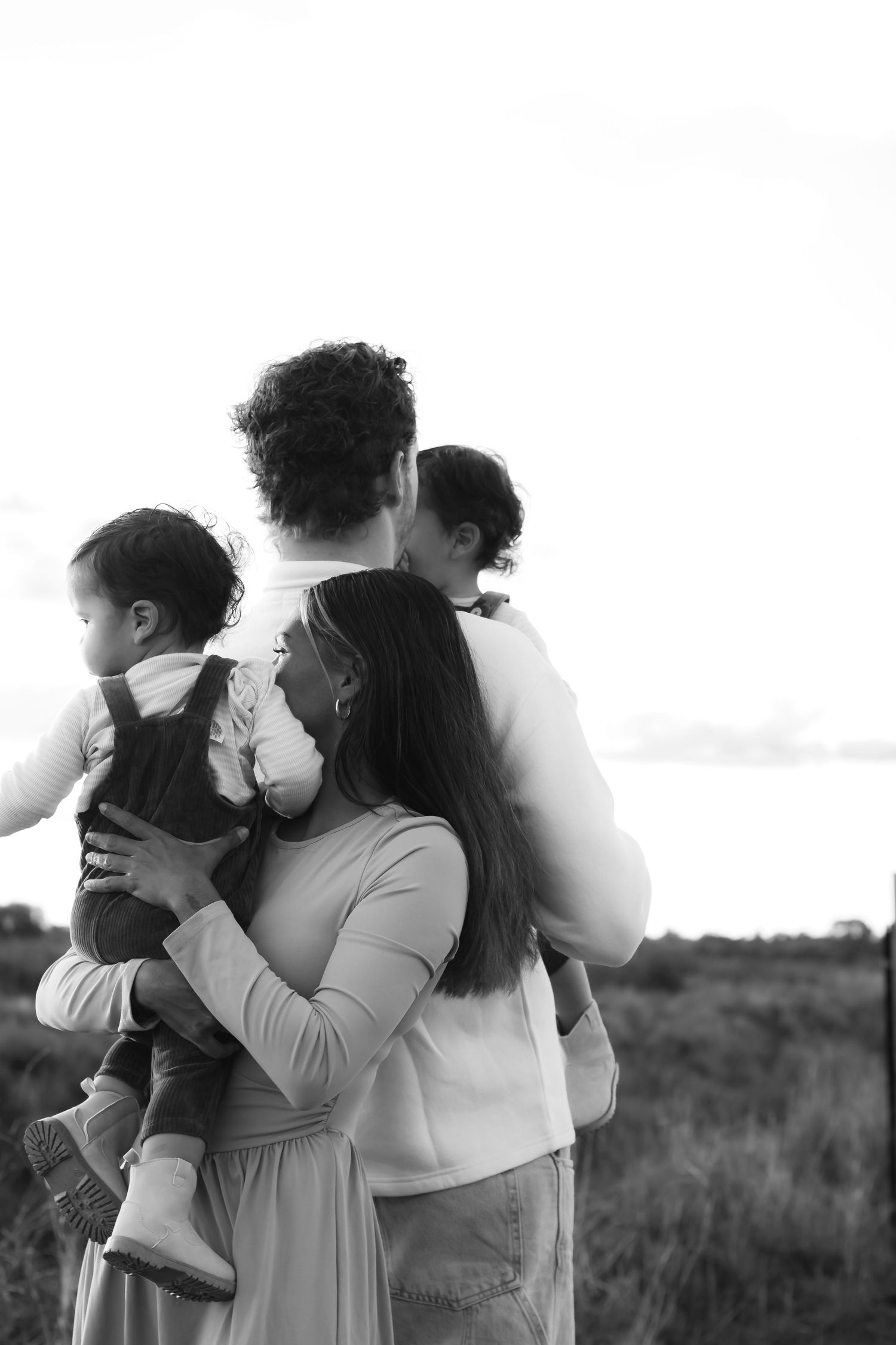 Black and white family portrait of parents holding two young children outdoors in a field at dusk.