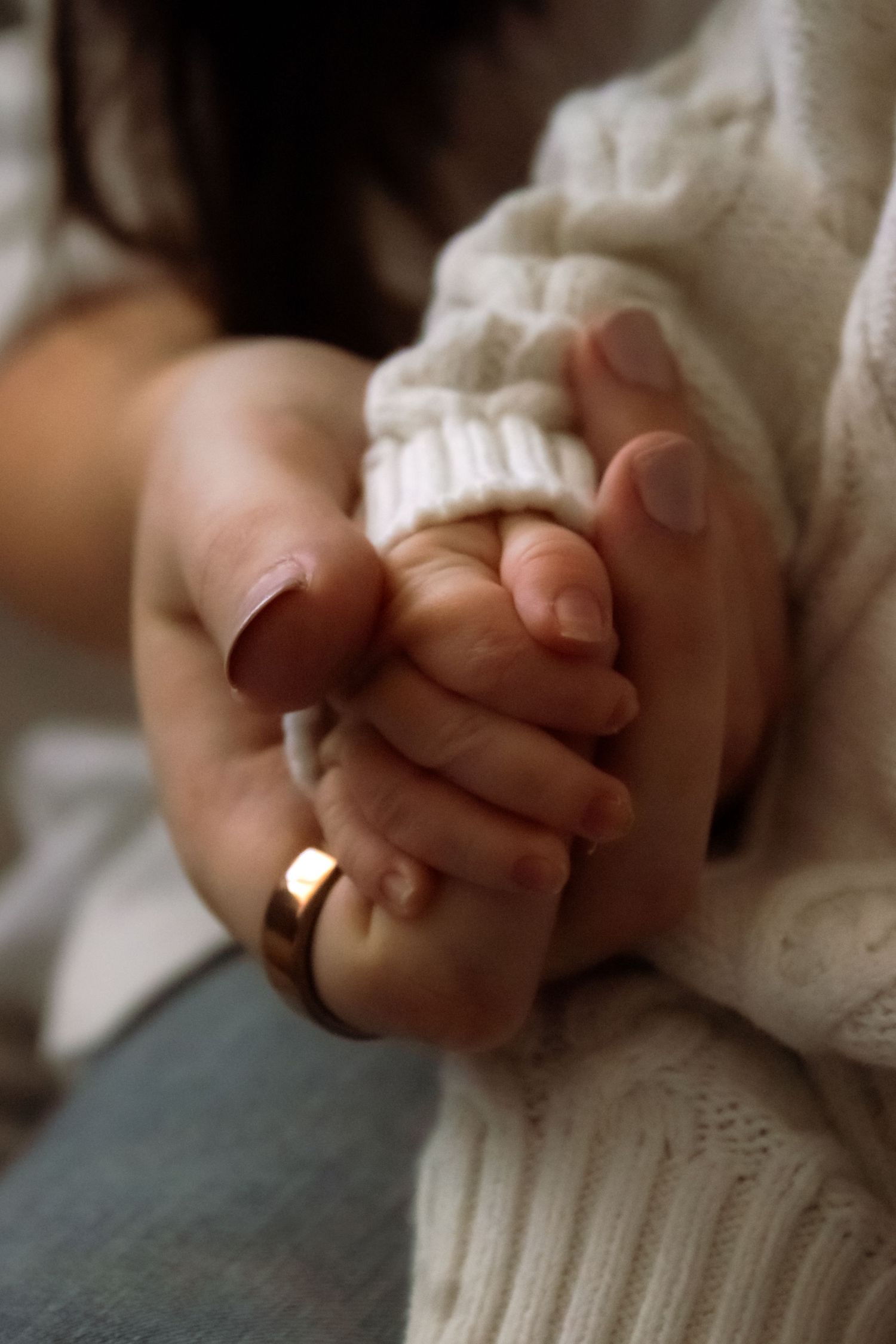 A mother's hand with gold wedding ring gently holds her newborn baby's tiny hand in a white knit sweater.