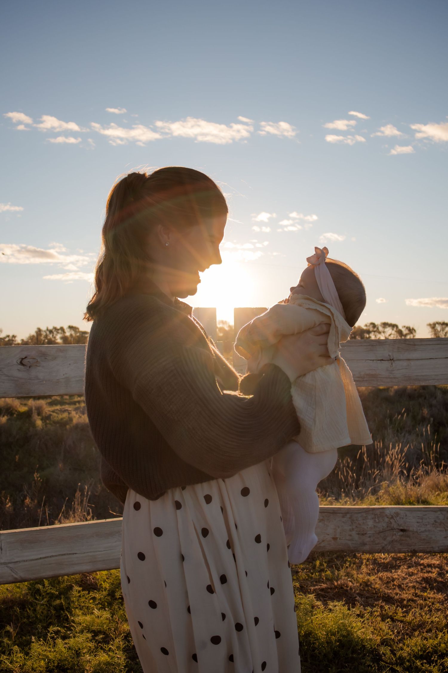 Mother holding newborn baby at golden sunset near white fence in rural countryside setting.