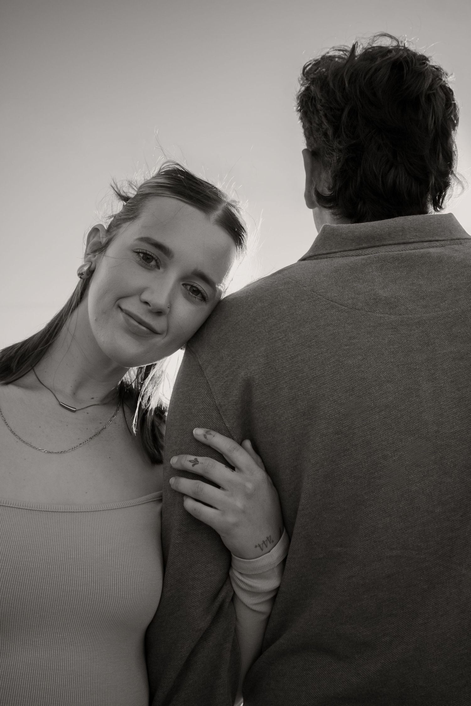 Black and white photo of a smiling young woman leaning on a mans shoulder outdoors.