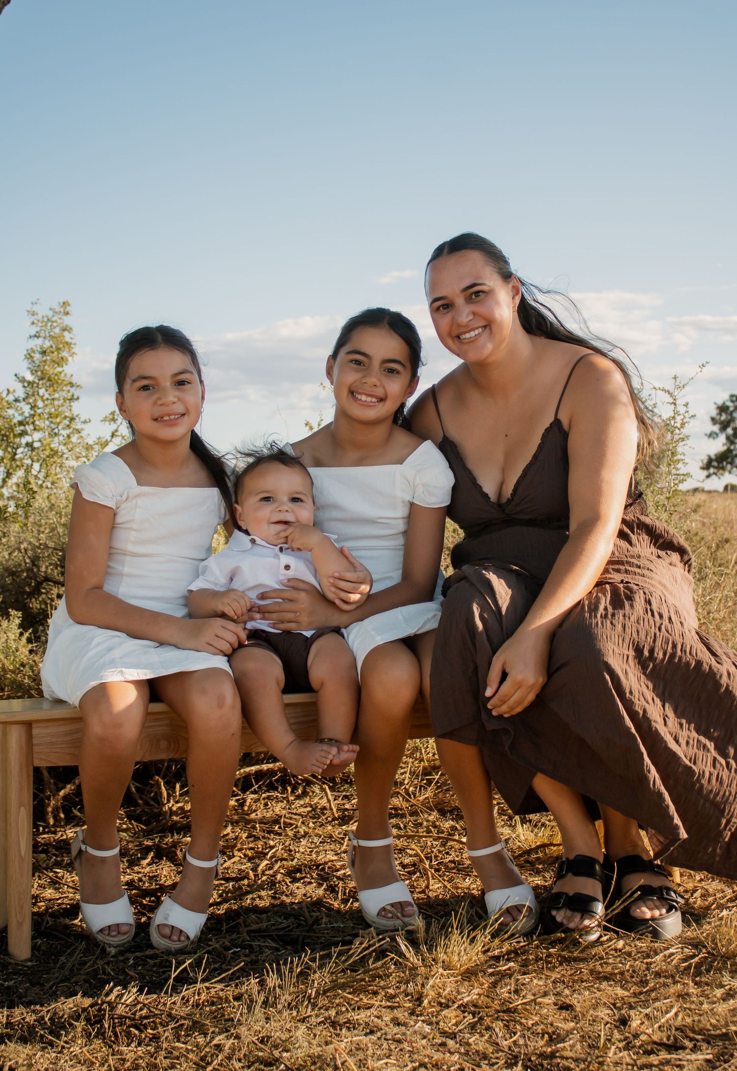 A mother and three children smile together outdoors on a sunny day, seated on a wooden bench in a natural setting.