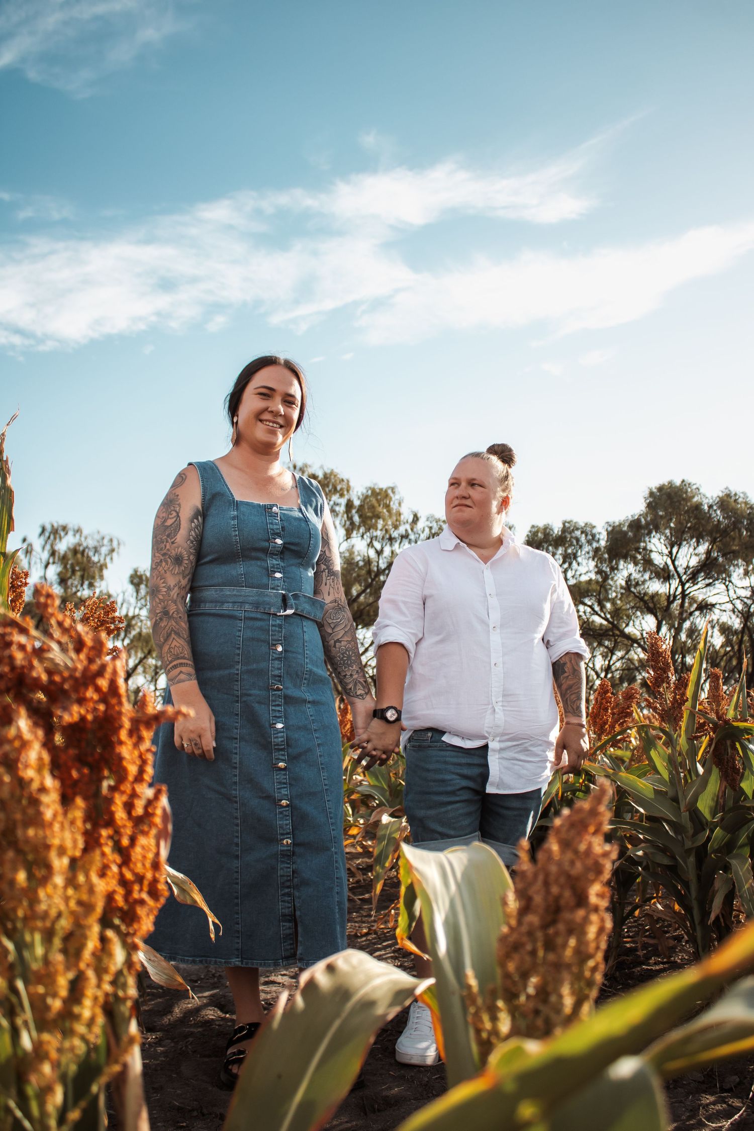 Couple holding hands in a sorghum field, woman in denim dress with tattoos, man in white shirt, blue sky background.