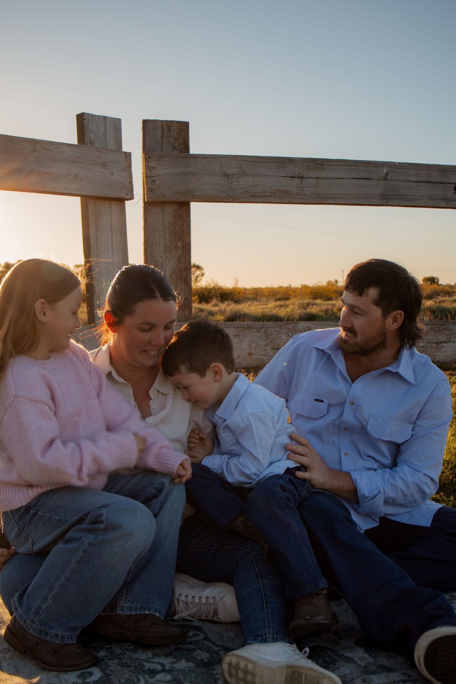 Family of four sitting together at golden hour near a wooden fence in an open rural landscape.
