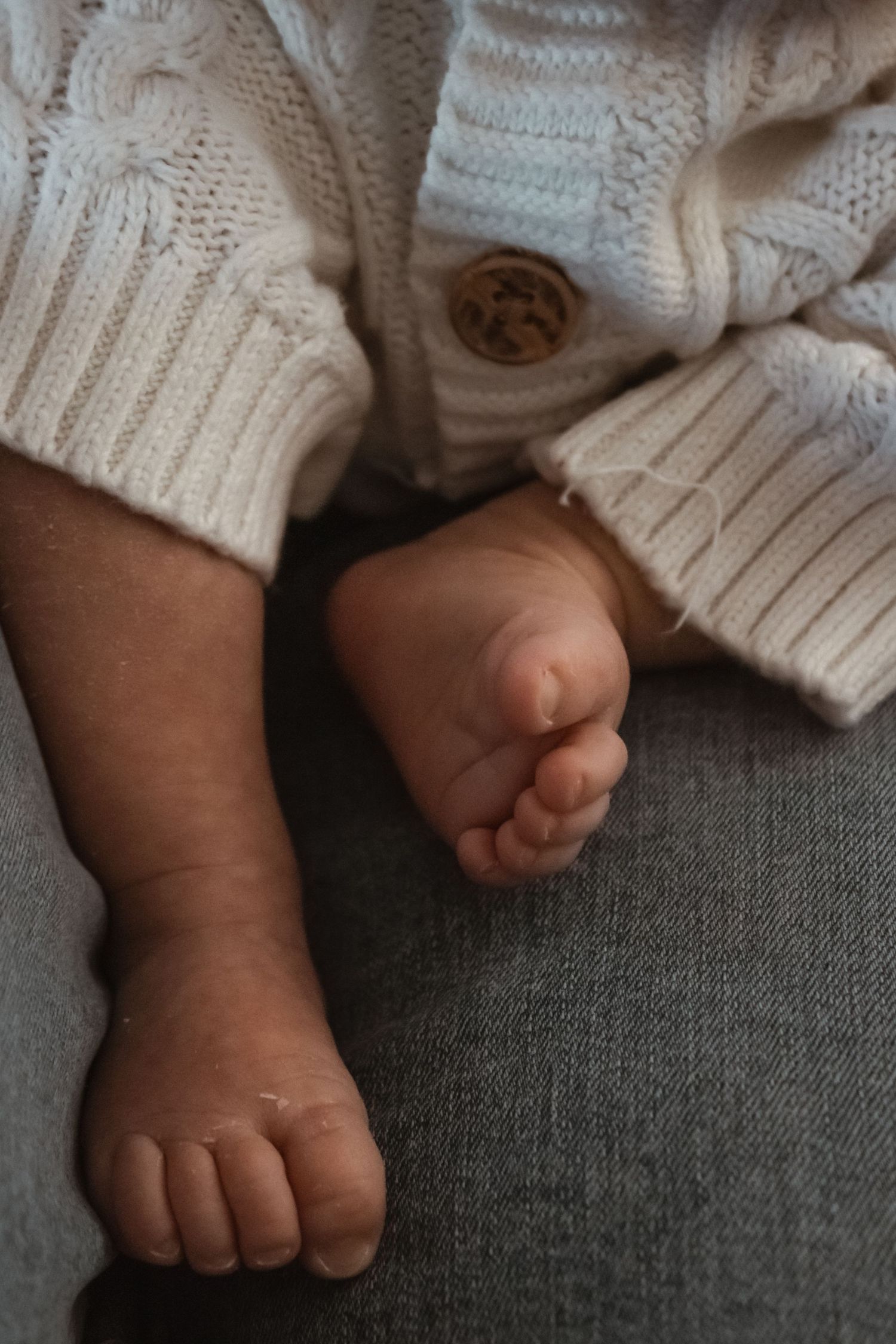 Close-up of tiny newborn baby feet wearing a white knit sweater, resting on gray fabric.