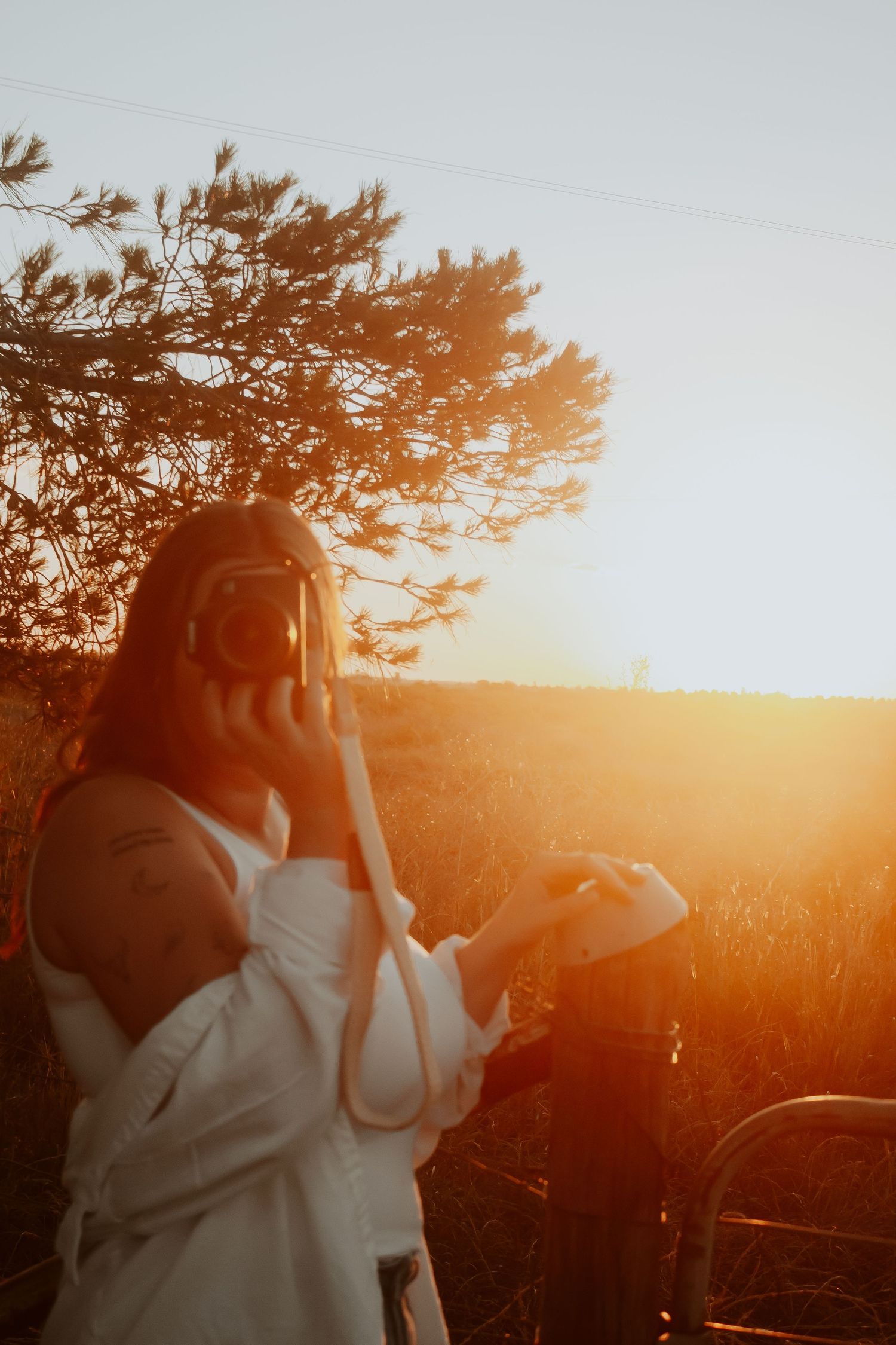 Woman in white shirt photographing with DSLR camera during golden sunset near pine tree in open field.