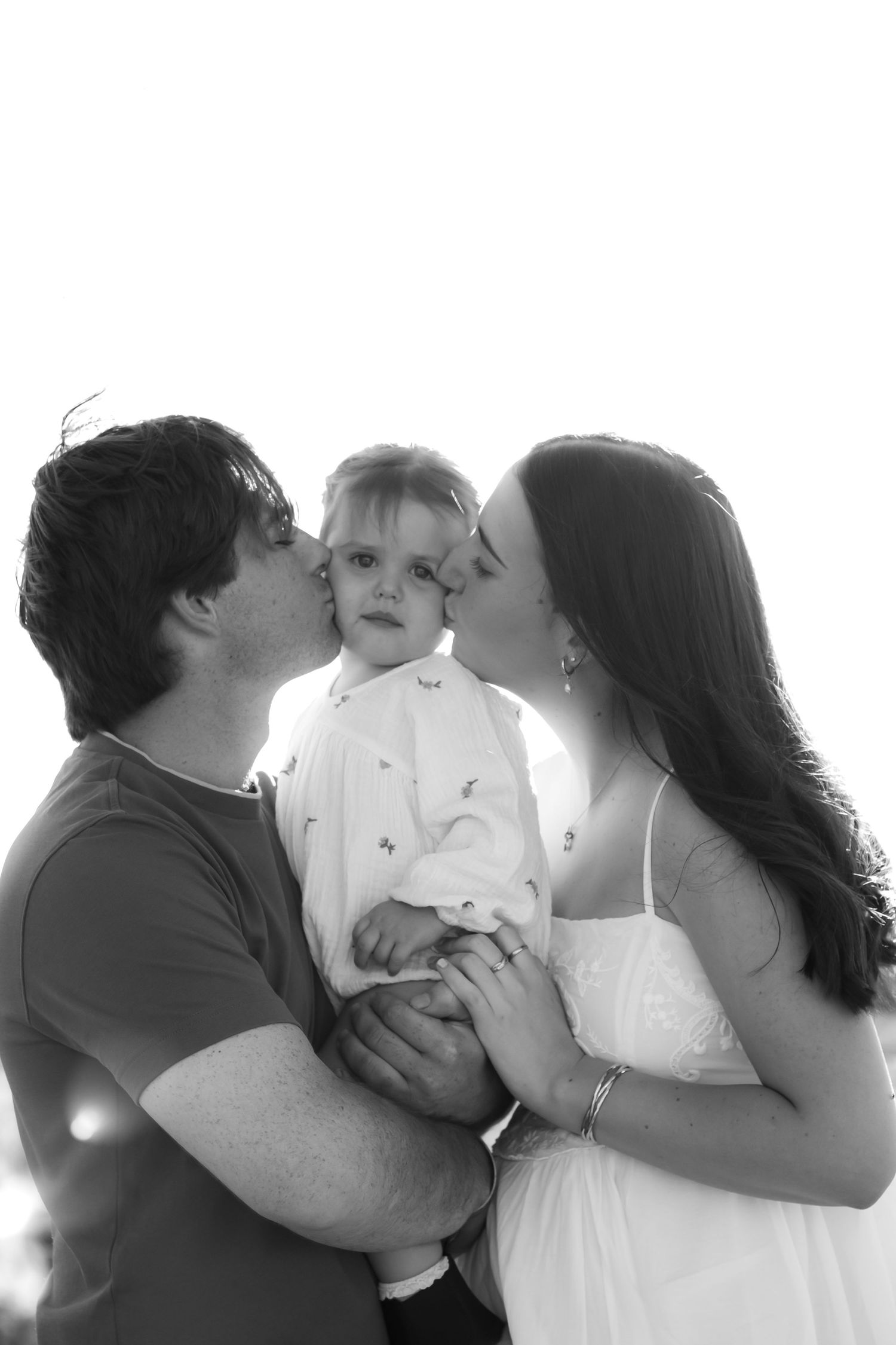 Black and white photo of parents kissing their toddler on the cheeks in a tender family portrait.