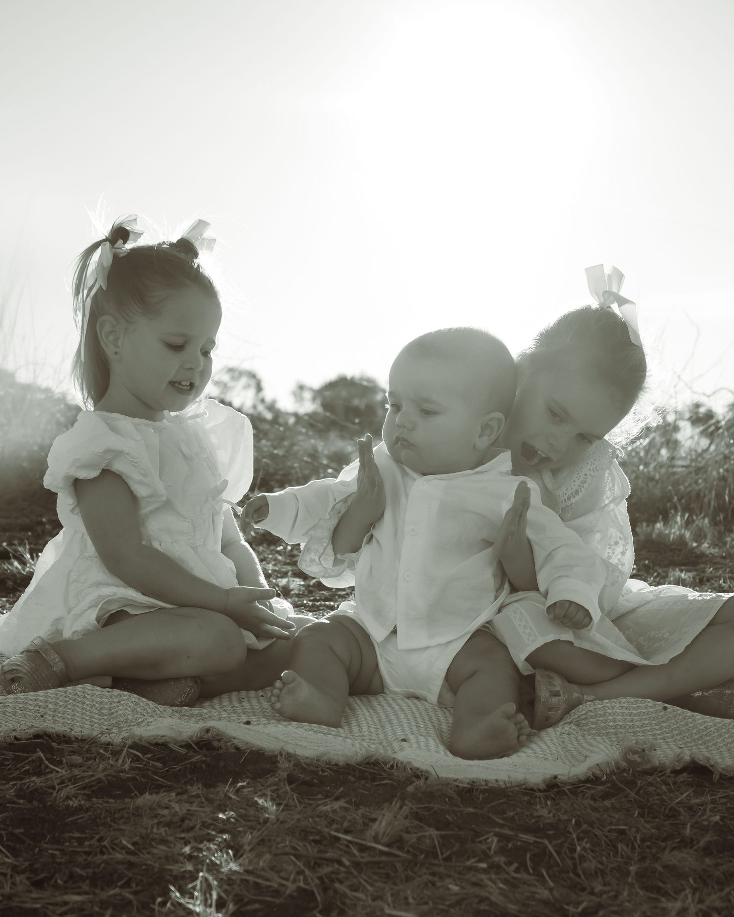 Three young children in white outfits sit together on a blanket outdoors in a charming black and white photo.