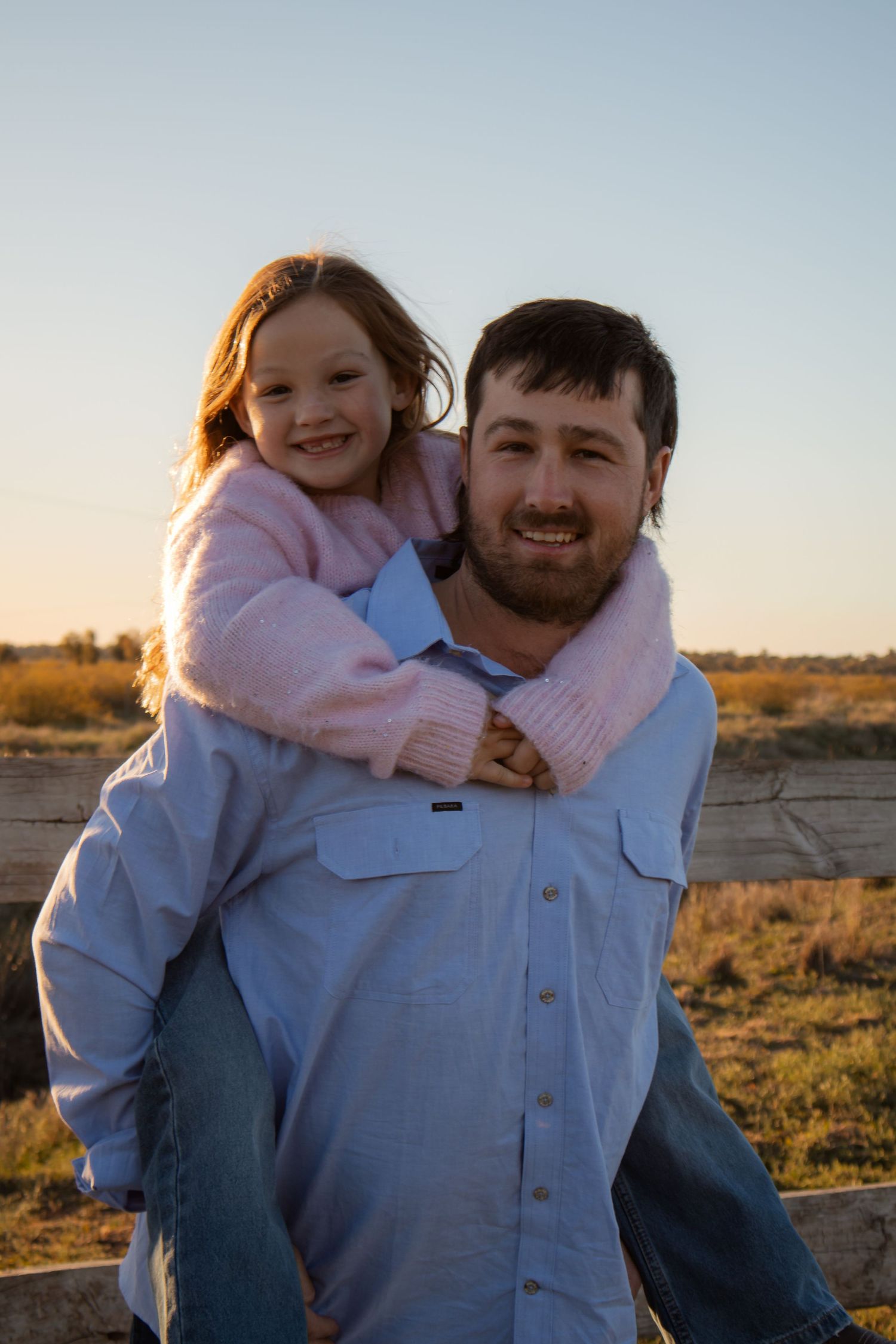 A smiling man giving a happy young girl a piggyback ride outdoors at golden hour near a wooden fence.