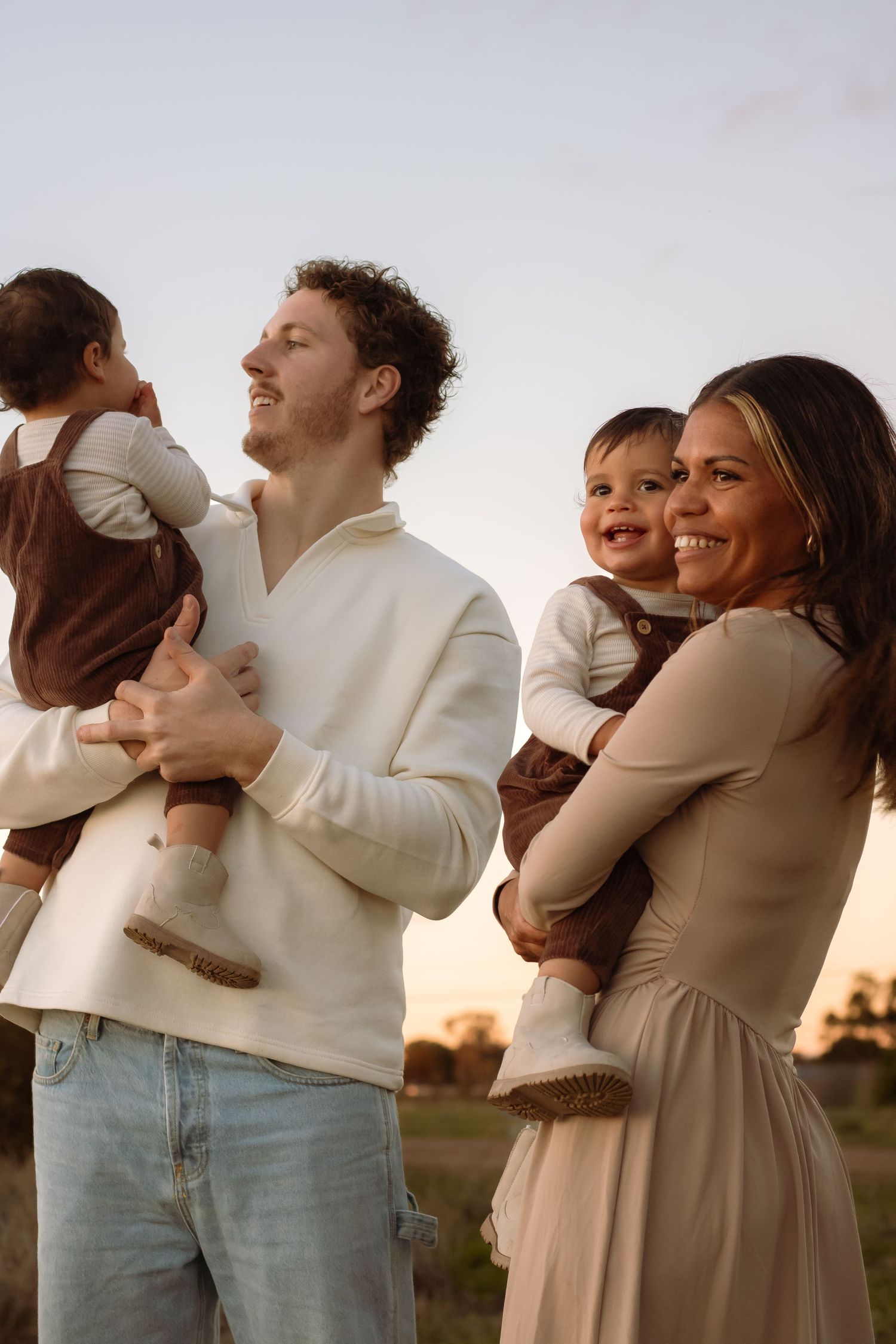 A happy young family with two babies poses outdoors at sunset, smiling warmly in neutral-toned outfits.