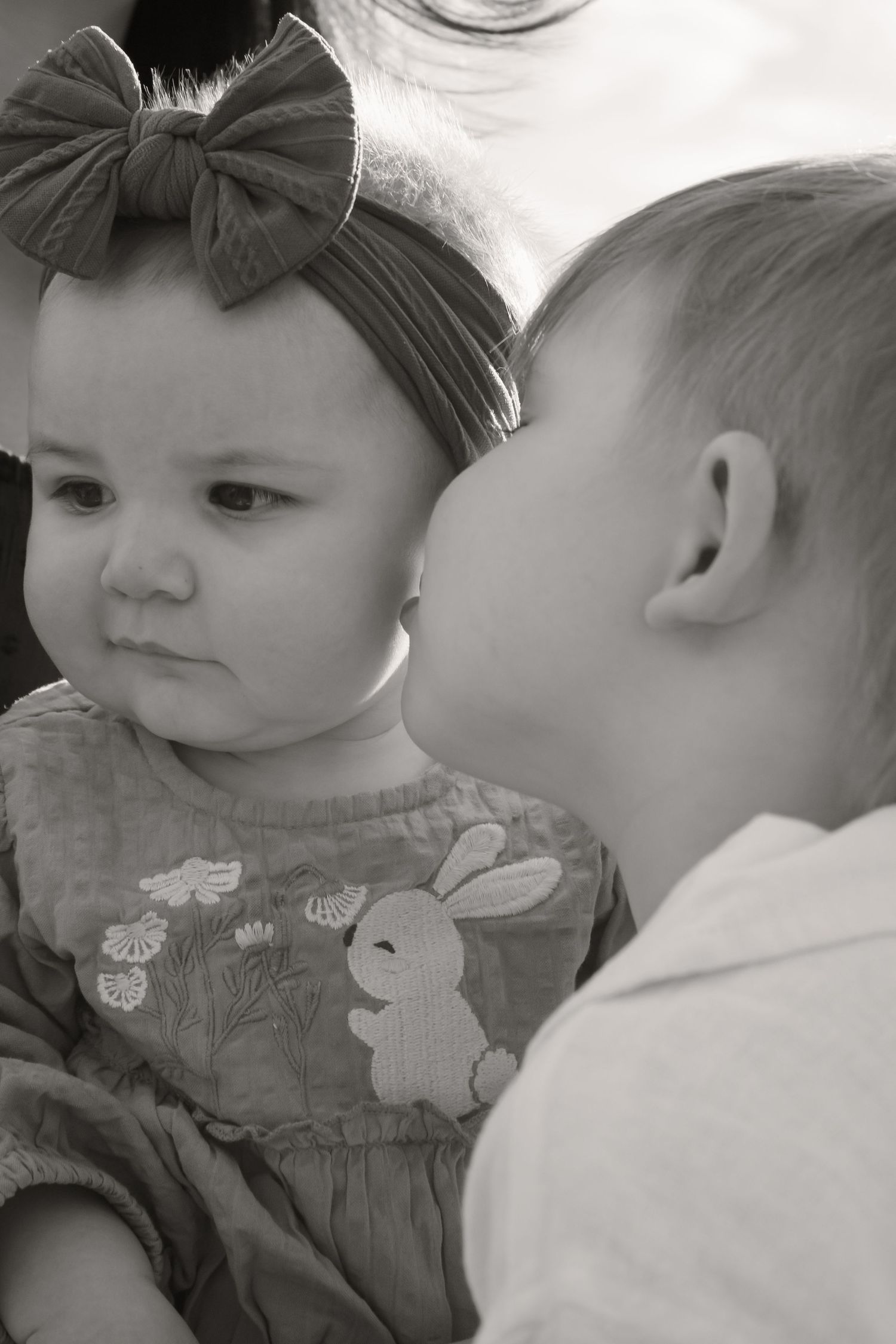 Black and white photo of a young boy gently kissing a baby girl wearing a bow headband and bunny dress.