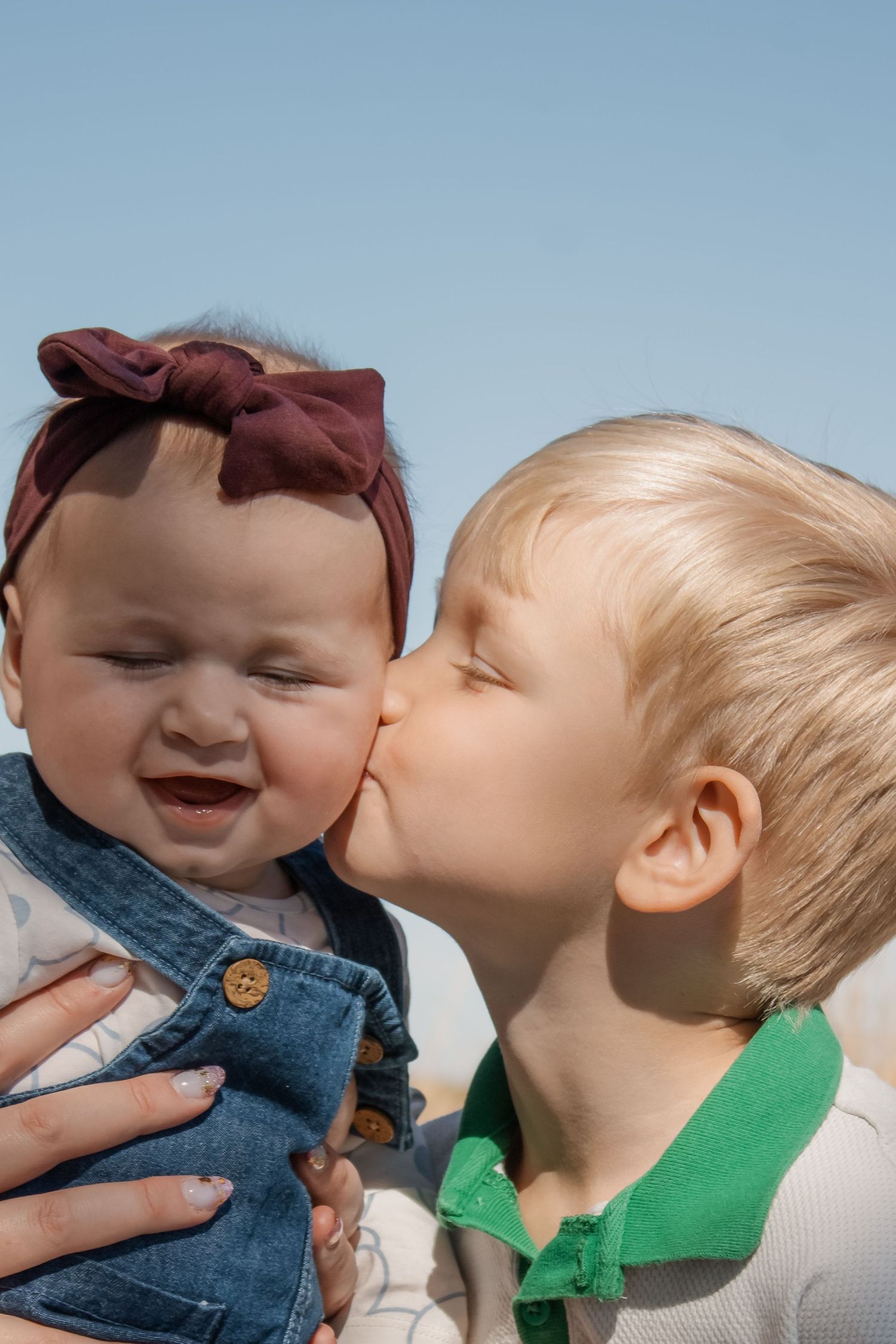 A young boy kisses a smiling baby girl wearing a maroon bow headband outdoors under a clear blue sky.