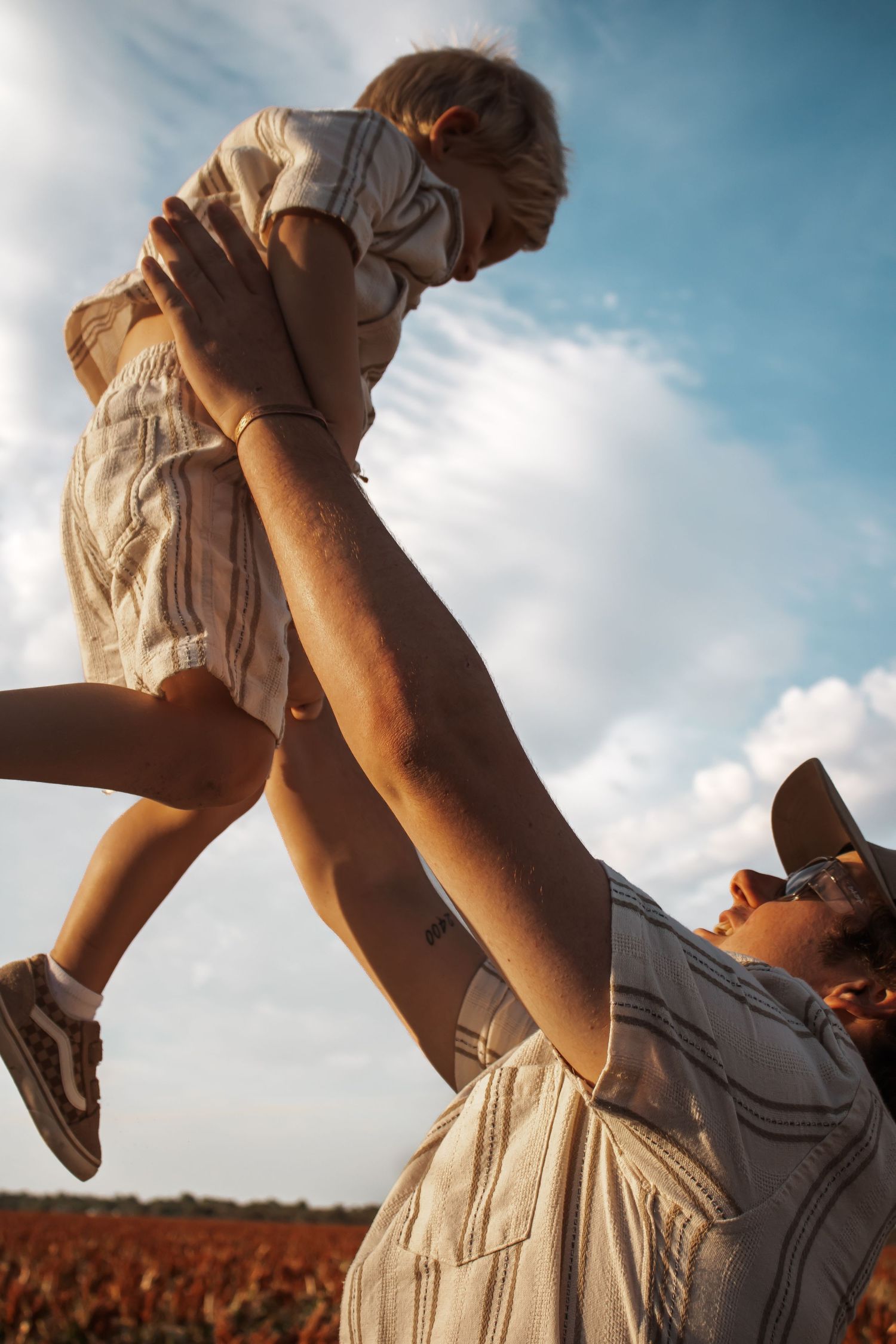 Parent joyfully lifting child into the sky in a sunlit field, both wearing casual linen outfits on a sunny day.
