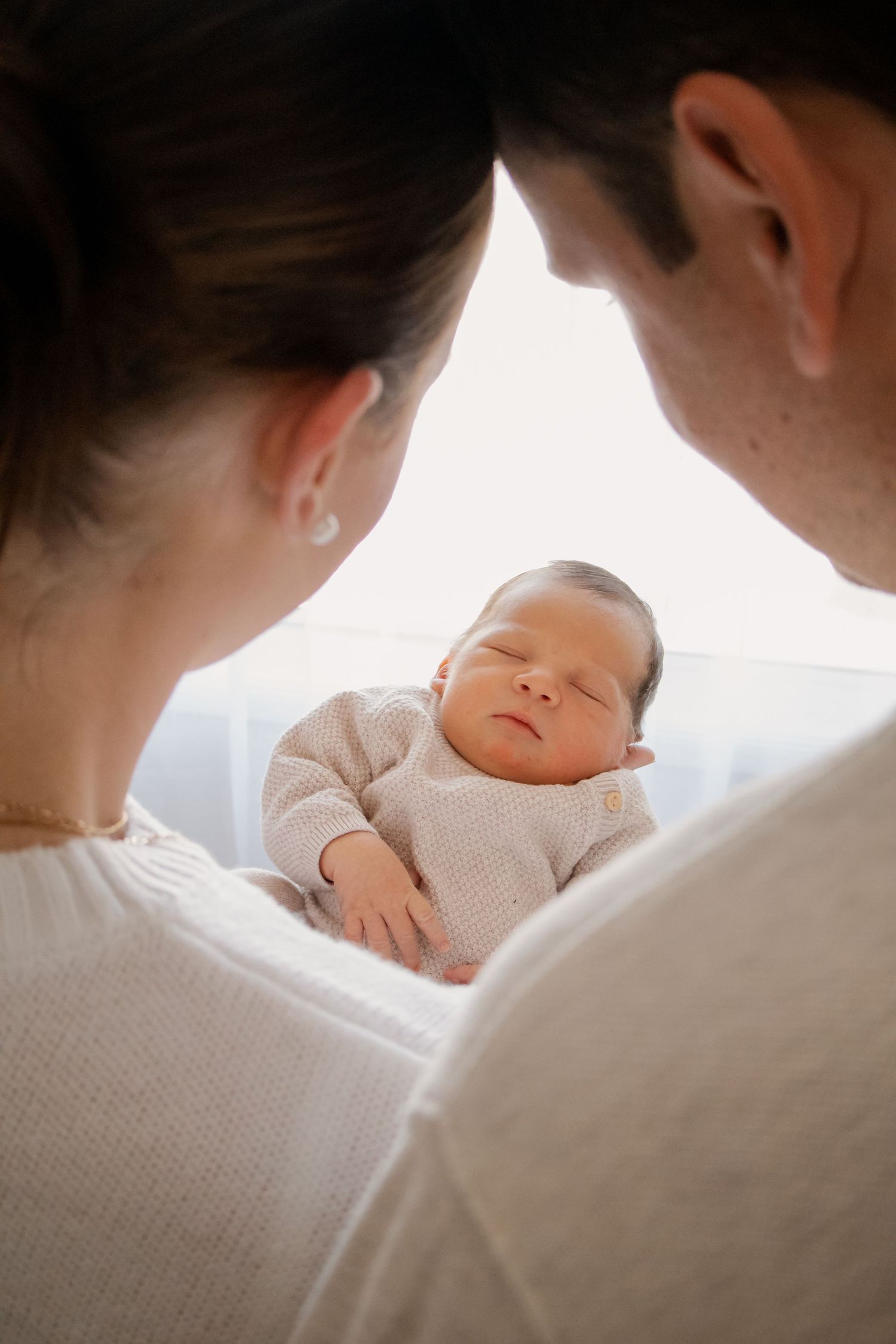 New parents gazing lovingly at their sleeping newborn baby dressed in a beige knit onesie.