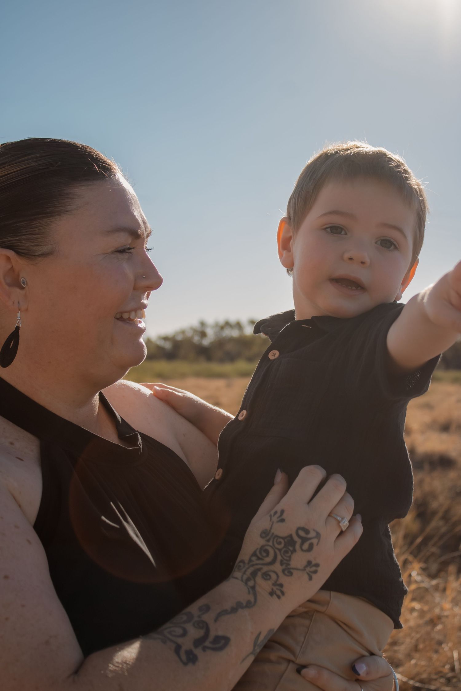 A smiling woman with floral tattoo holds a young boy outdoors in a sunny field.