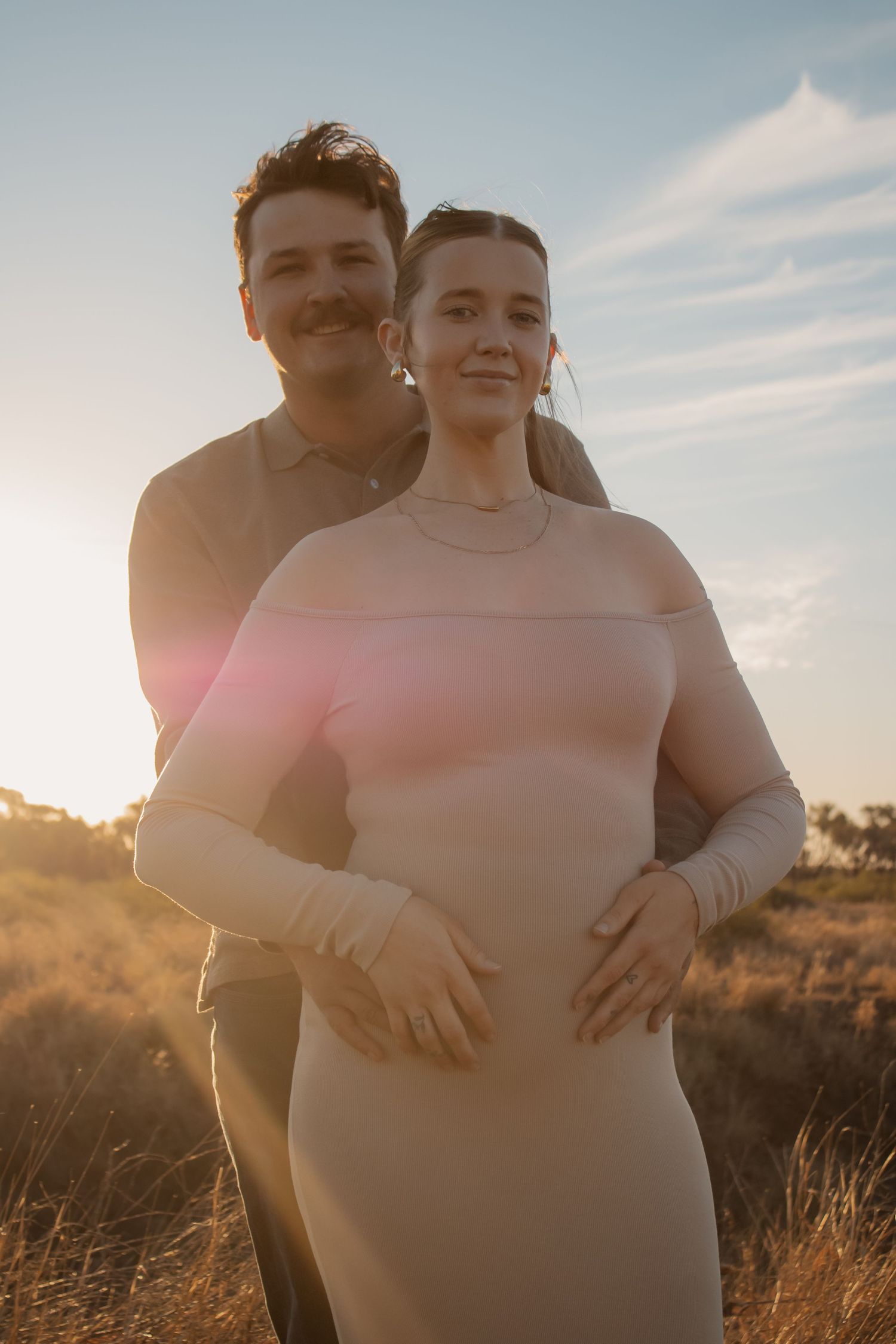 Couple embracing at golden hour sunset in a field, woman in pink dress, man in olive shirt, lens flare.