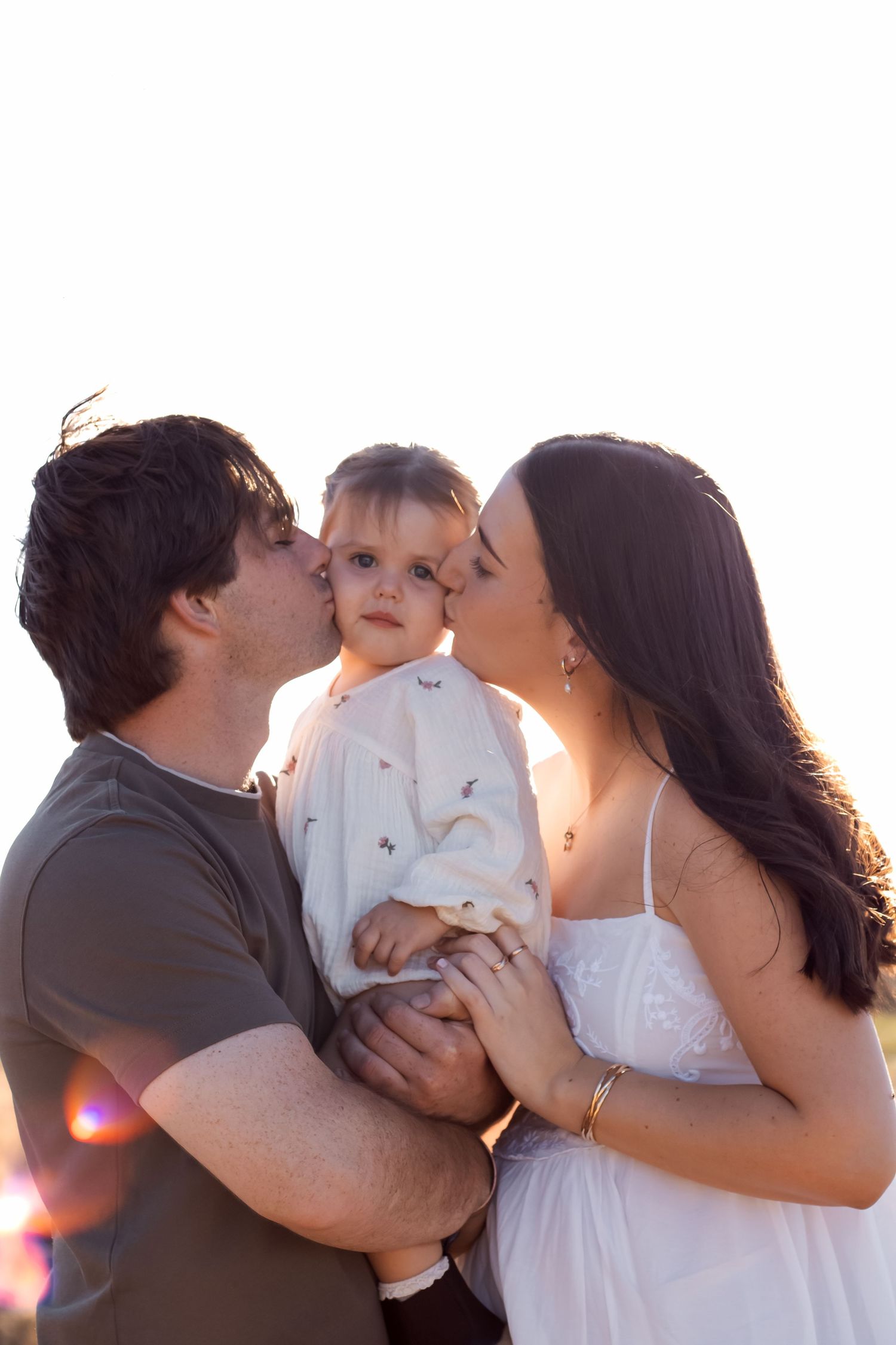 Parents kissing their baby on the cheeks outdoors at sunset in a heartwarming family portrait.