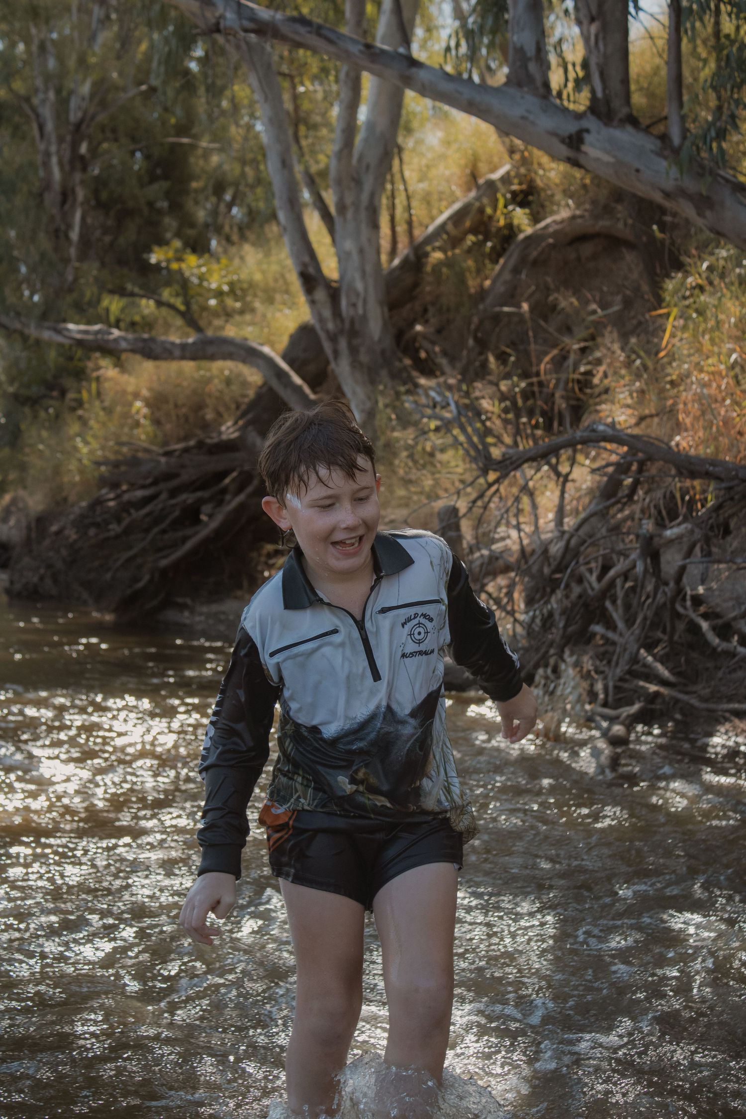 A smiling boy wades through a shallow creek surrounded by fallen trees and bush in an outdoor Australian setting.