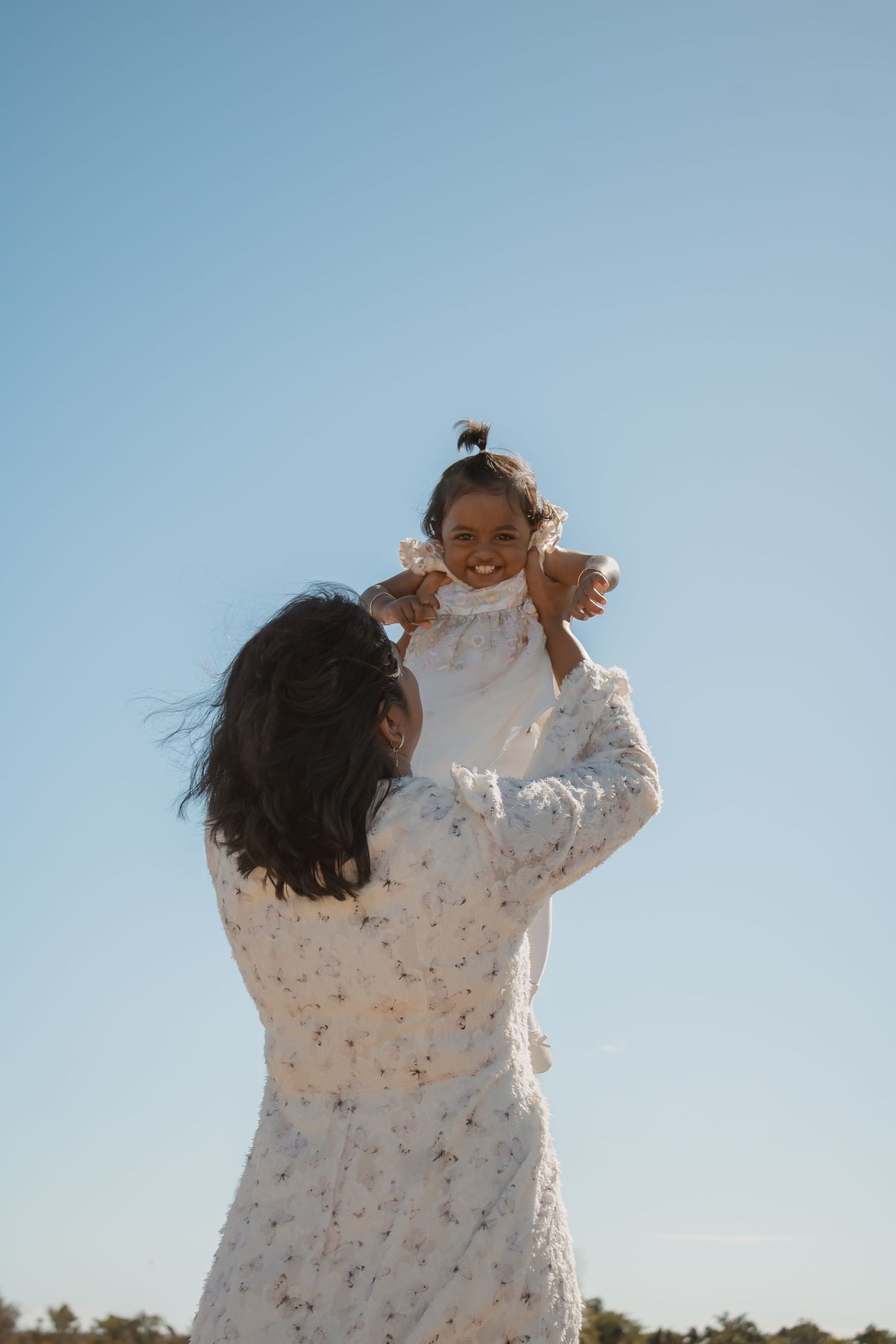 A mother in a floral dress lifts her smiling toddler high against a clear blue sky outdoors.