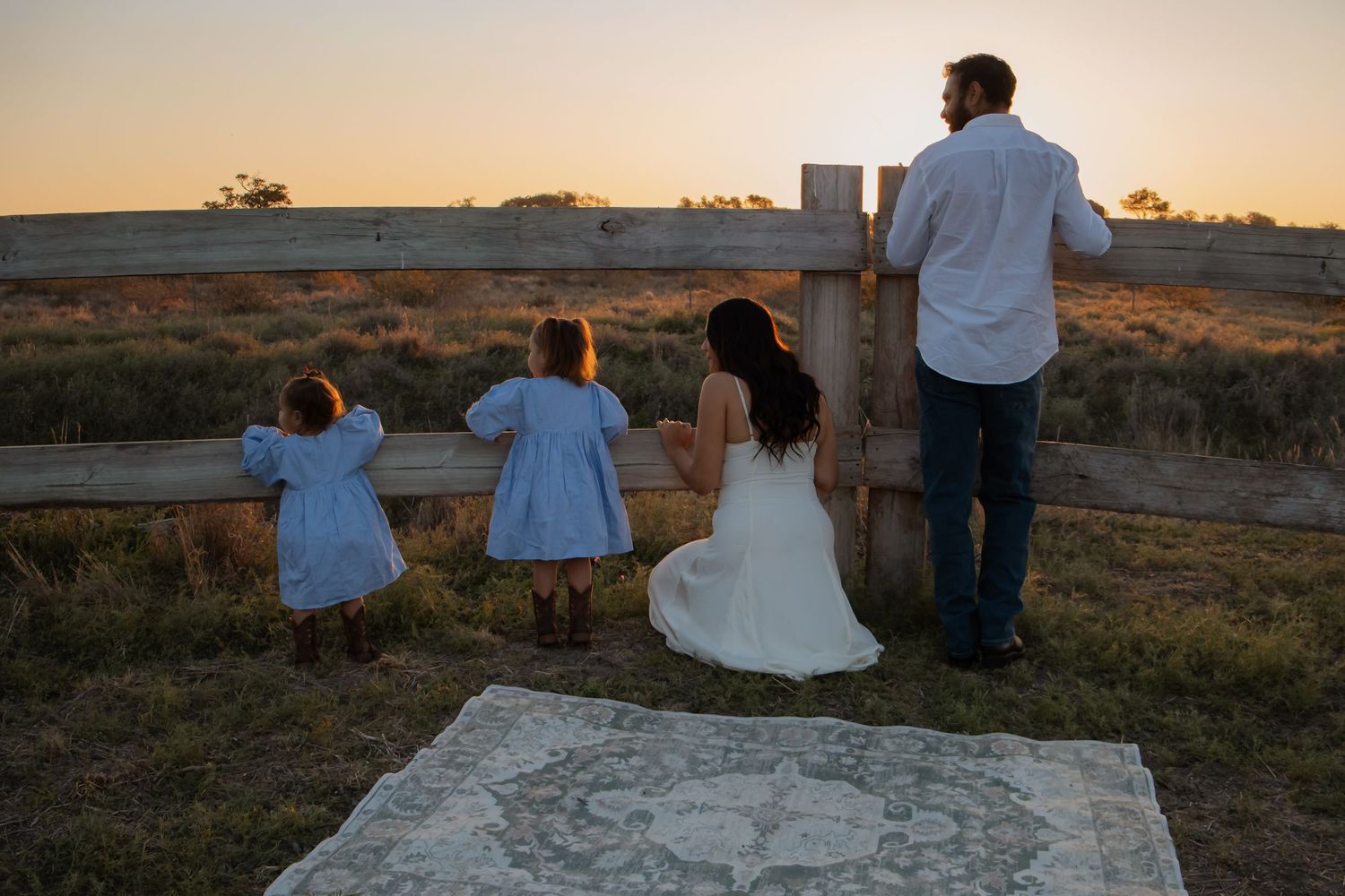 Family of four watching sunset at rustic fence on golden fields, bride in white gown with two young girls in blue dresses.