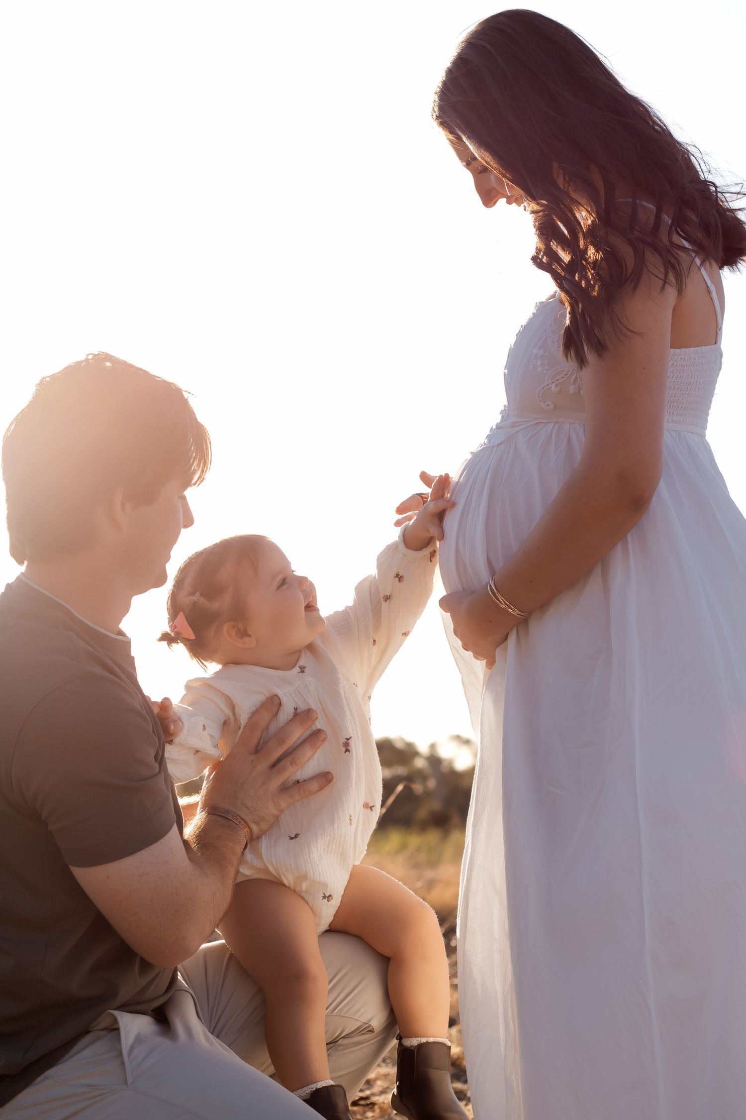 A pregnant woman in a white dress with her partner and toddler during a golden hour maternity photoshoot outdoors.