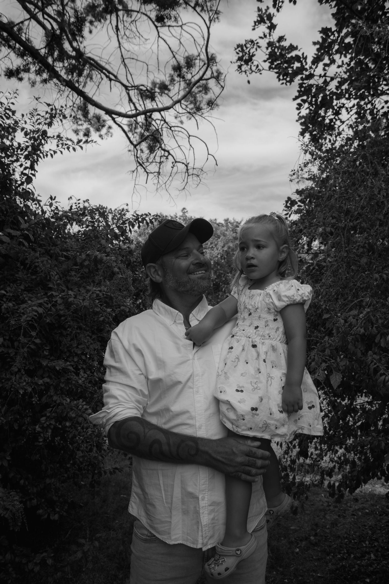 Black and white photo of a man holding a young girl outdoors, surrounded by trees and cloudy sky.