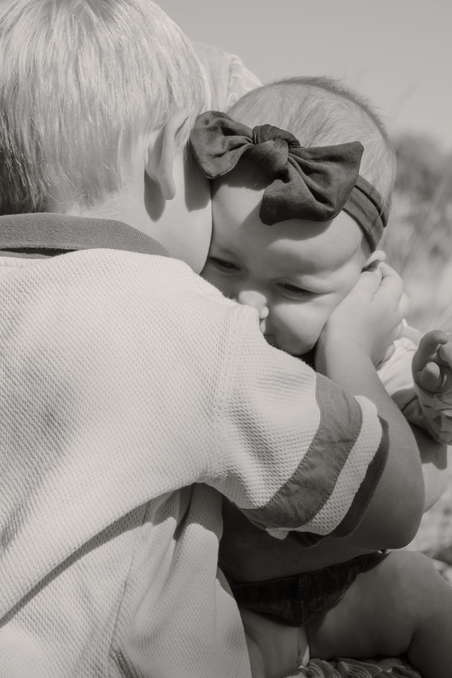 Black and white photo of a young boy hugging a baby girl wearing a bow headband outdoors.