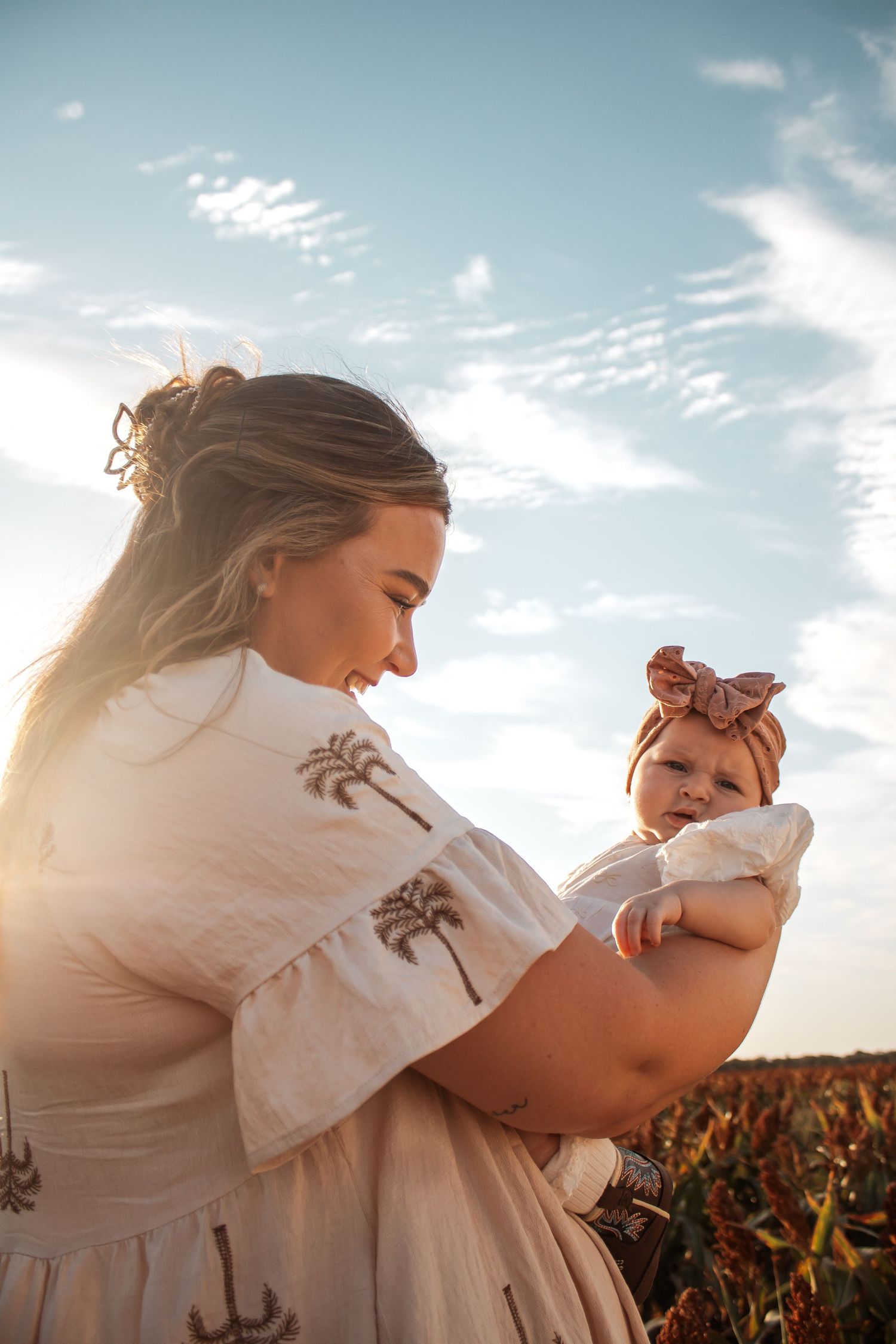 A mother holds her baby outdoors at golden hour, both wearing white, with a blue sky backdrop.