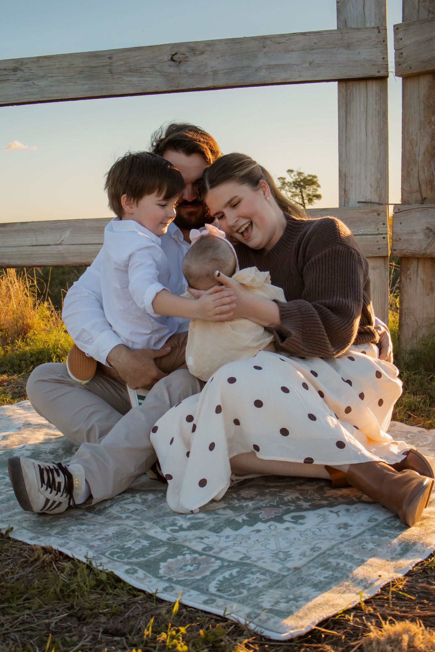 Happy family sitting on a blanket outdoors at sunset, parents laughing with two young children near a wooden fence.