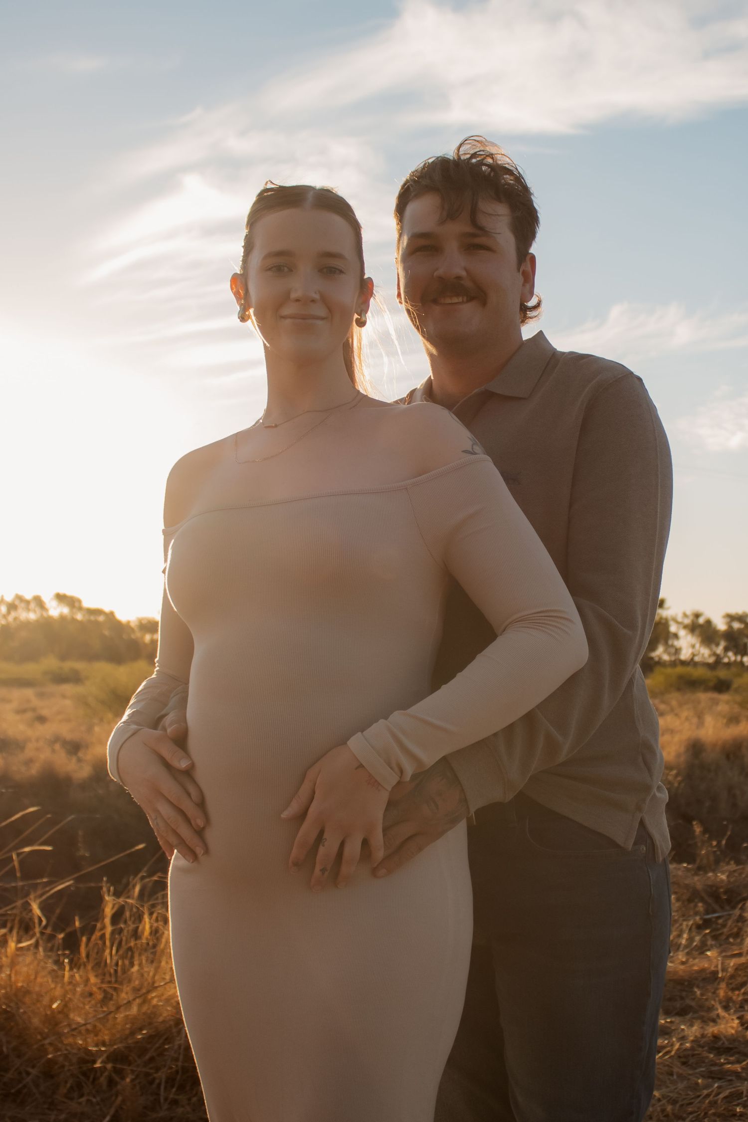 Couple poses outdoors at golden hour, woman in beige off-shoulder dress with partner embracing her from behind in field.