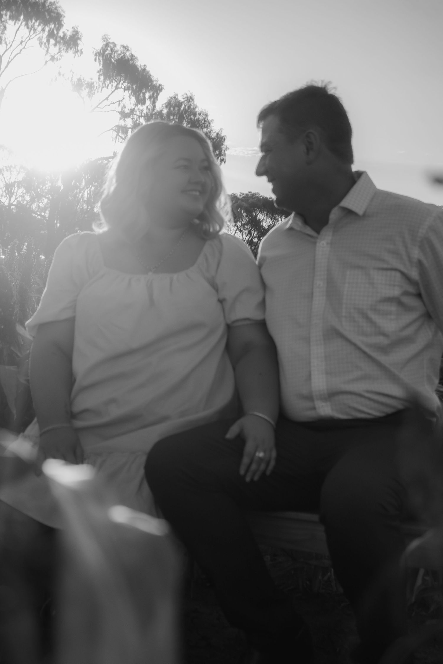 Black and white photo of a smiling couple sitting together outdoors, sharing a joyful moment in soft backlit sunlight.