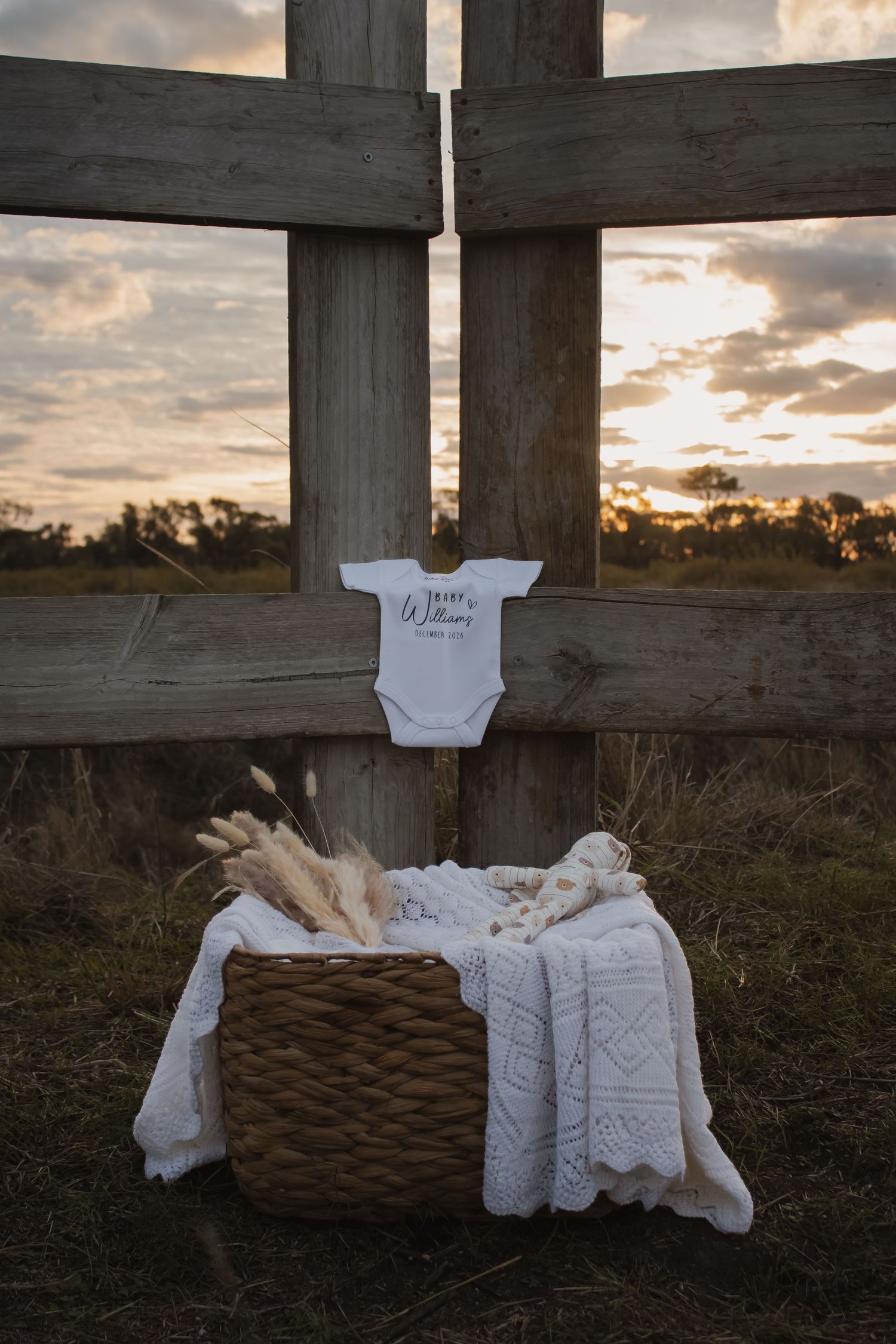 Baby onesie hanging on rustic wooden cross at sunset with wicker basket and white lace blanket below.