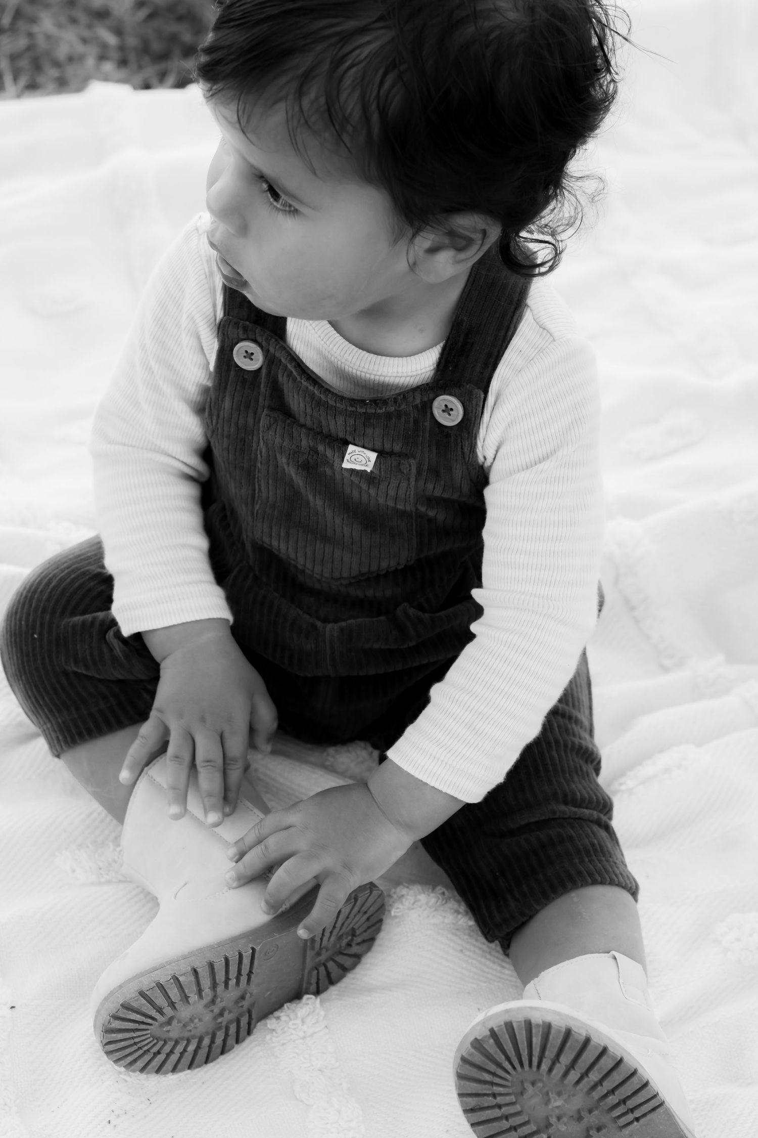Black and white photo of a toddler in overalls sitting on a blanket, looking to the side.