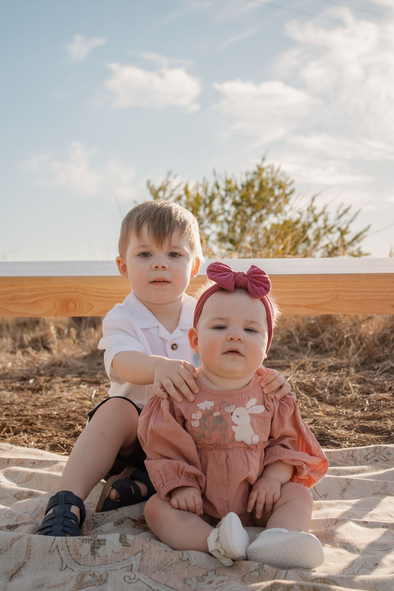 Two young siblings, a toddler boy and baby girl with pink bow, sitting together outdoors on a blanket in warm sunlight.