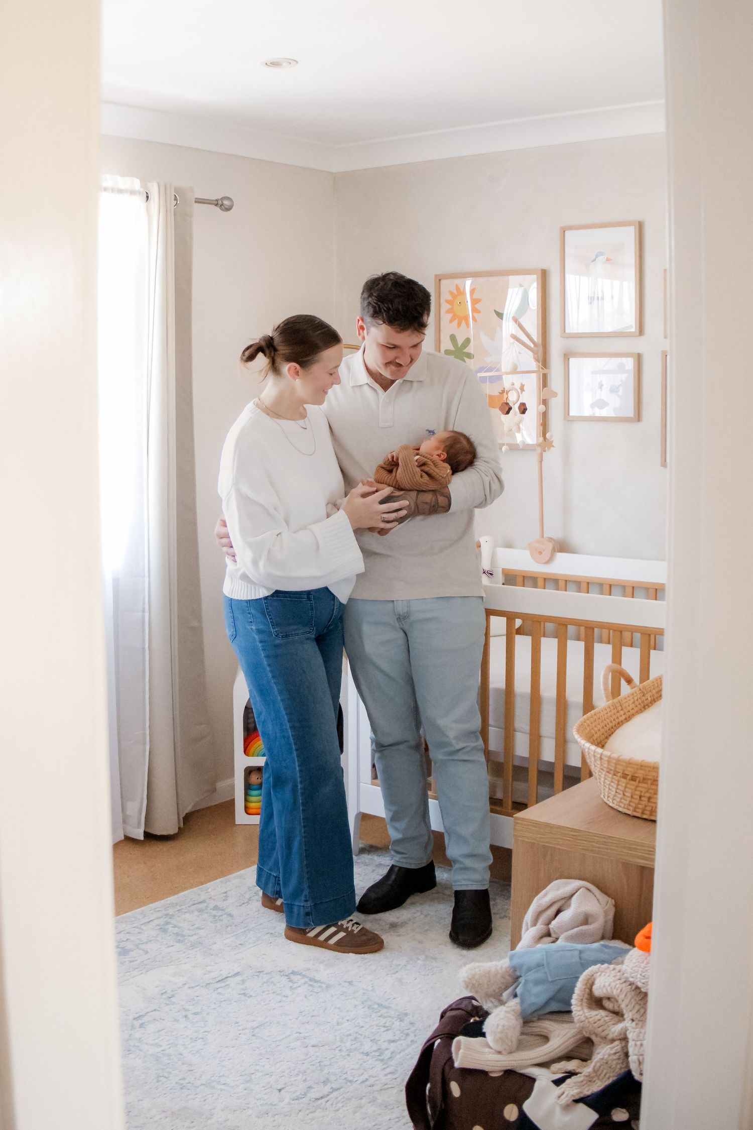 New parents stand together in a softly decorated nursery, lovingly cradling their newborn baby near a wooden crib.