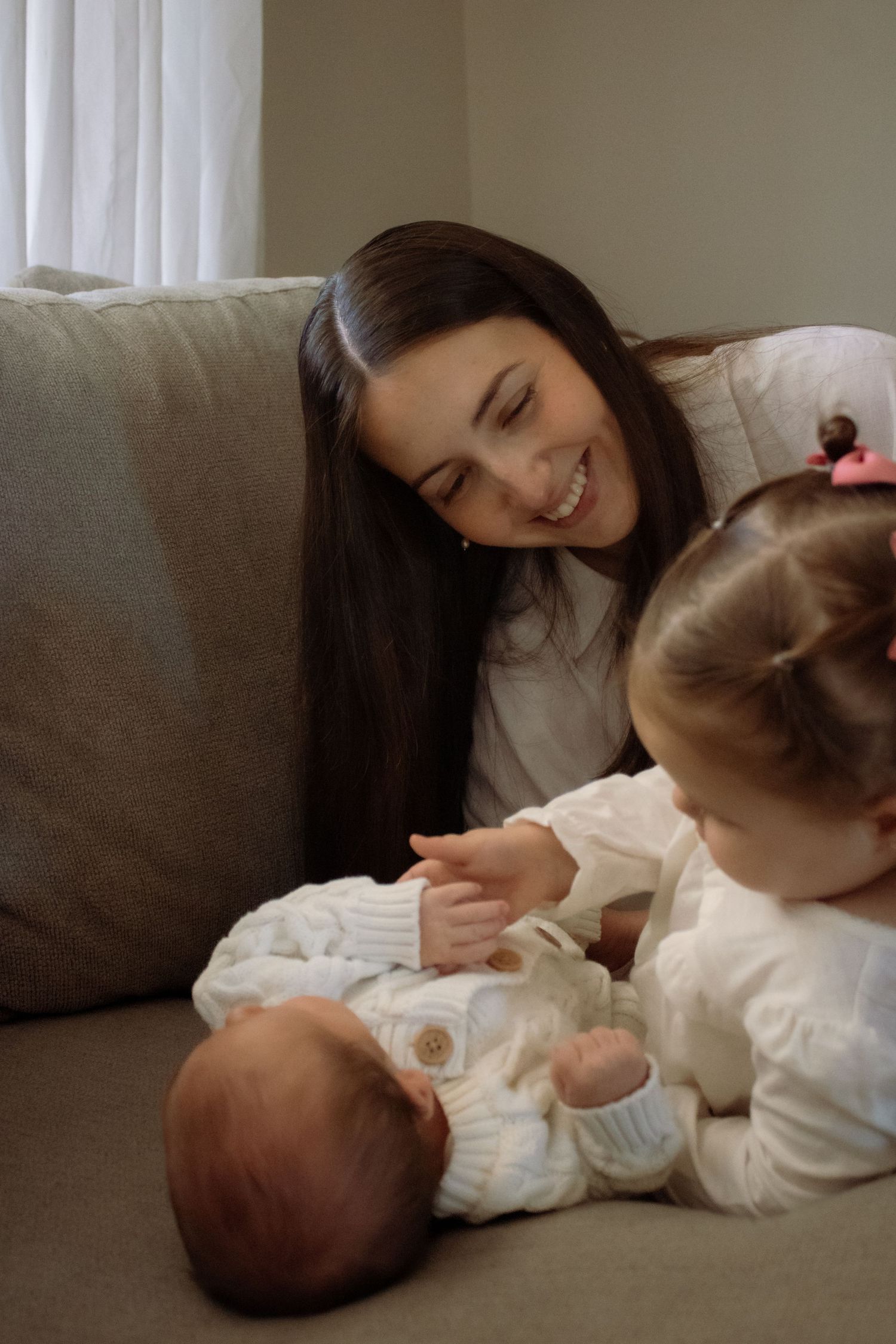 Smiling mother on sofa watching toddler interact with newborn baby, both children dressed in white.