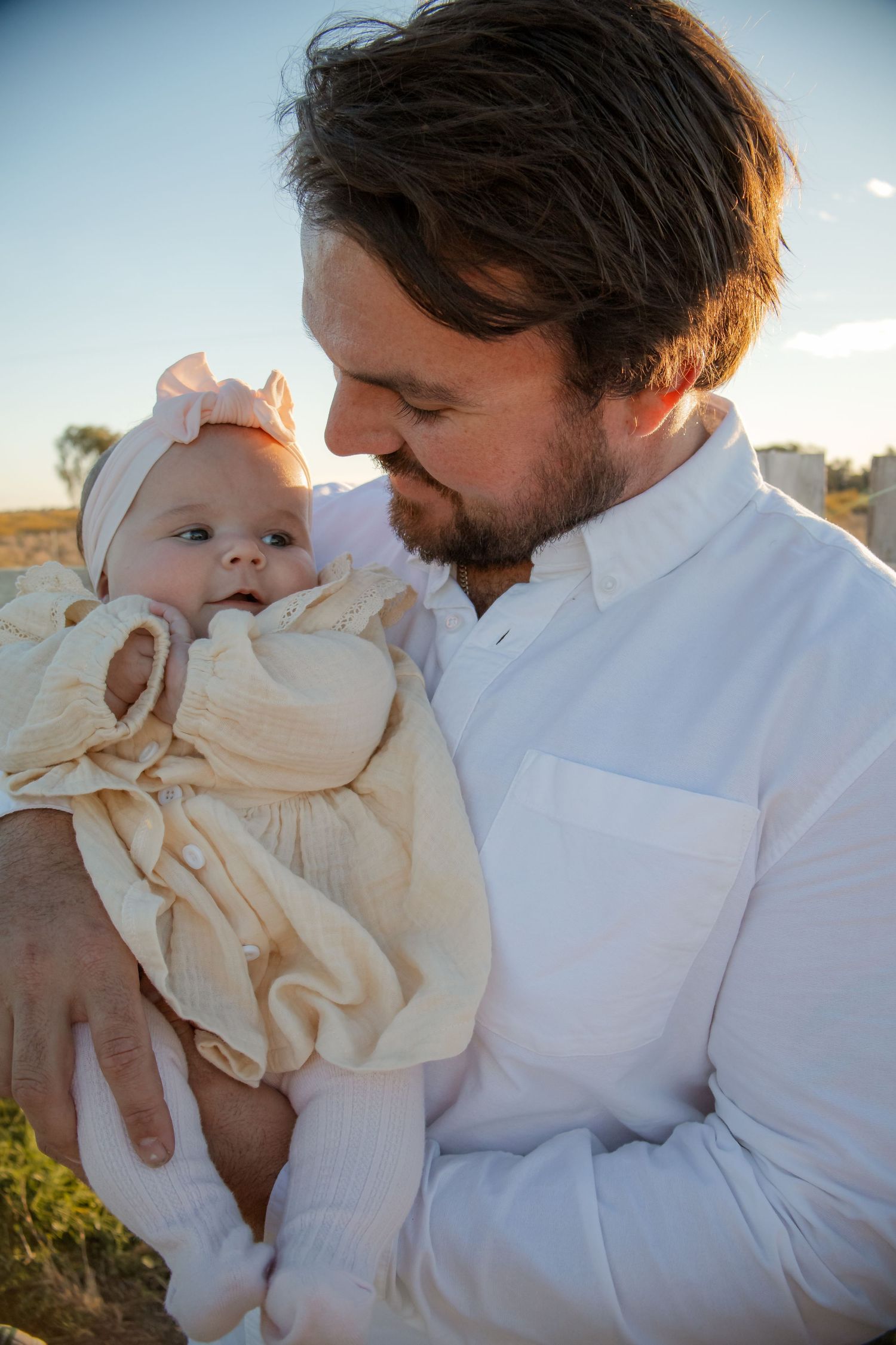 A bearded man in a white shirt tenderly holds a baby girl in a cream dress and pink bow outdoors.