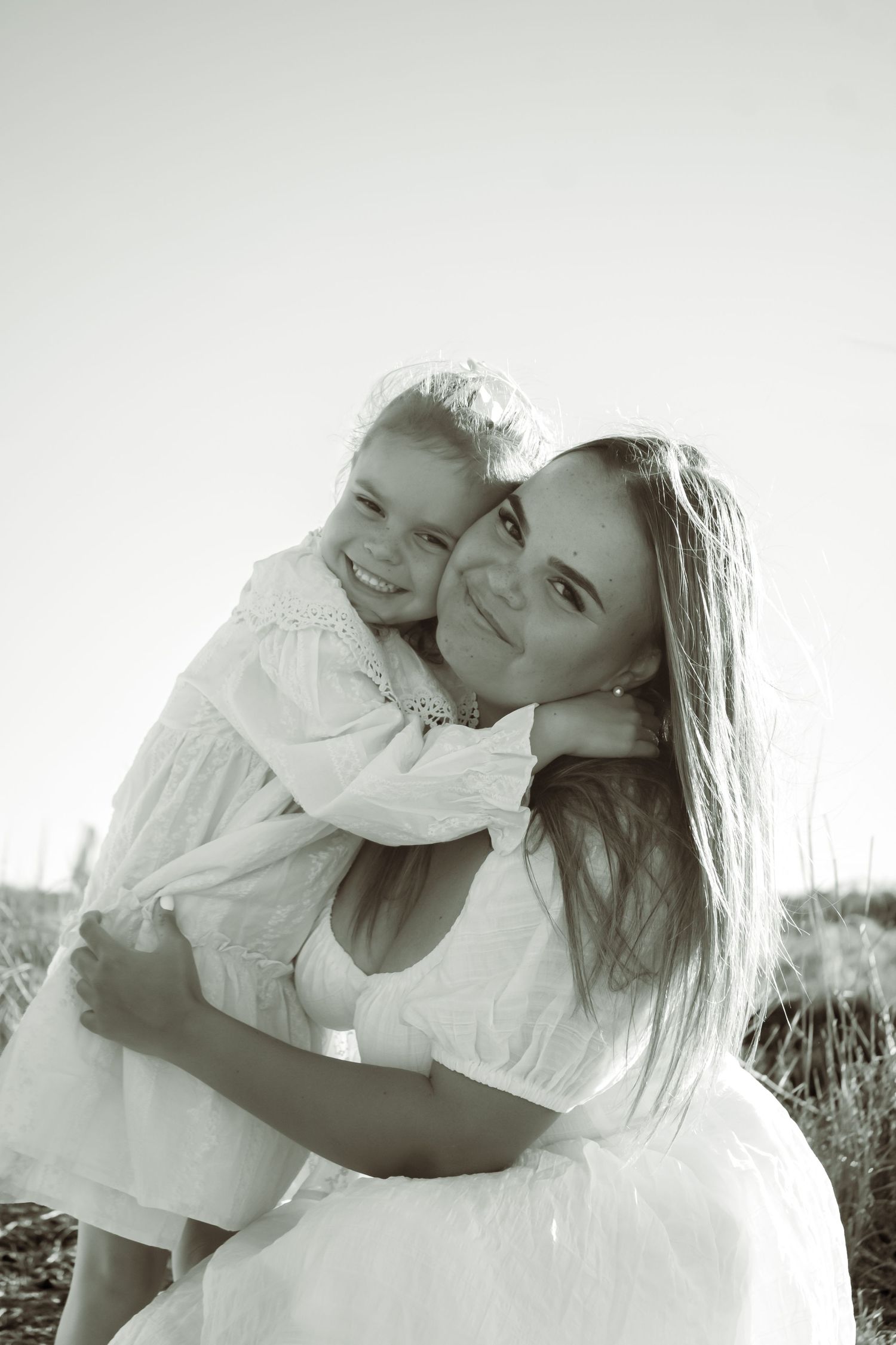 Black and white photo of a smiling mother hugging her young daughter in matching white dresses outdoors.