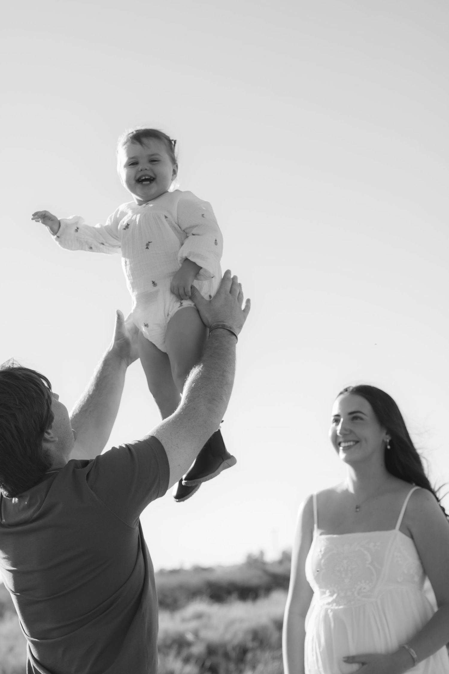 Black and white photo of a father tossing laughing baby in the air while pregnant mother watches and smiles outdoors.