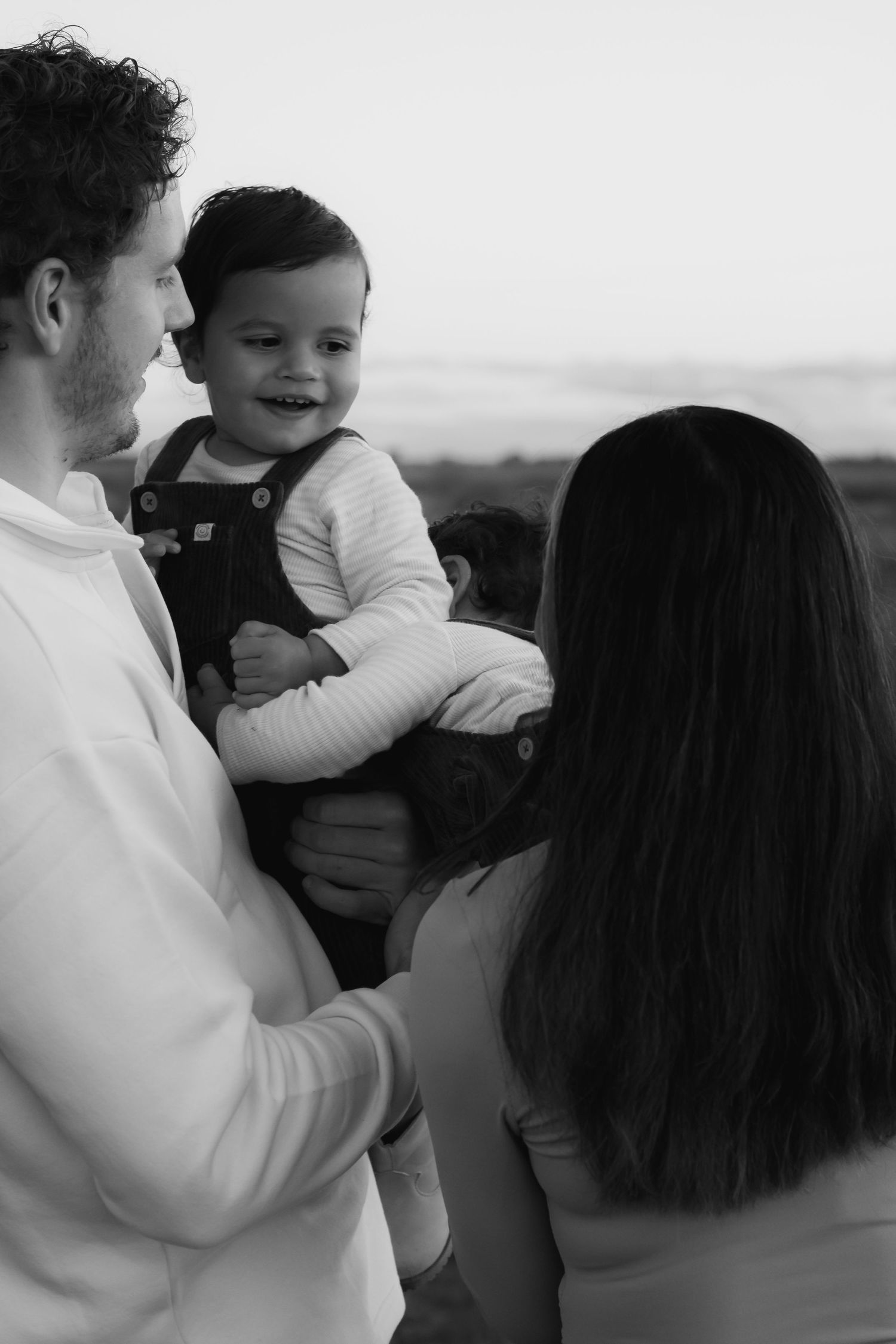 Black and white photo of a smiling baby held by father while mother looks on outdoors.