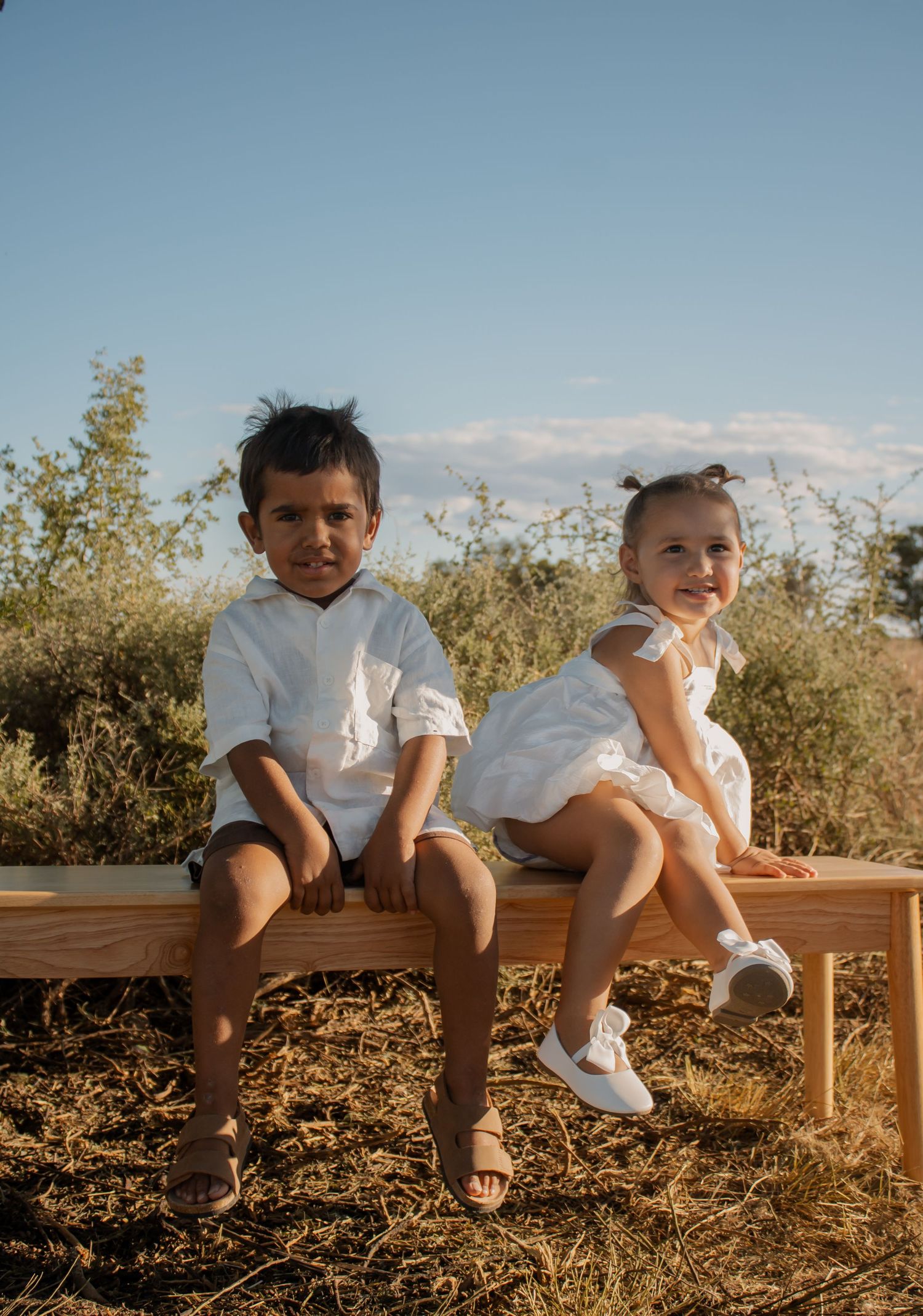 Two young children in white outfits sit on a wooden bench outdoors, smiling, with shrubs and blue sky behind them.