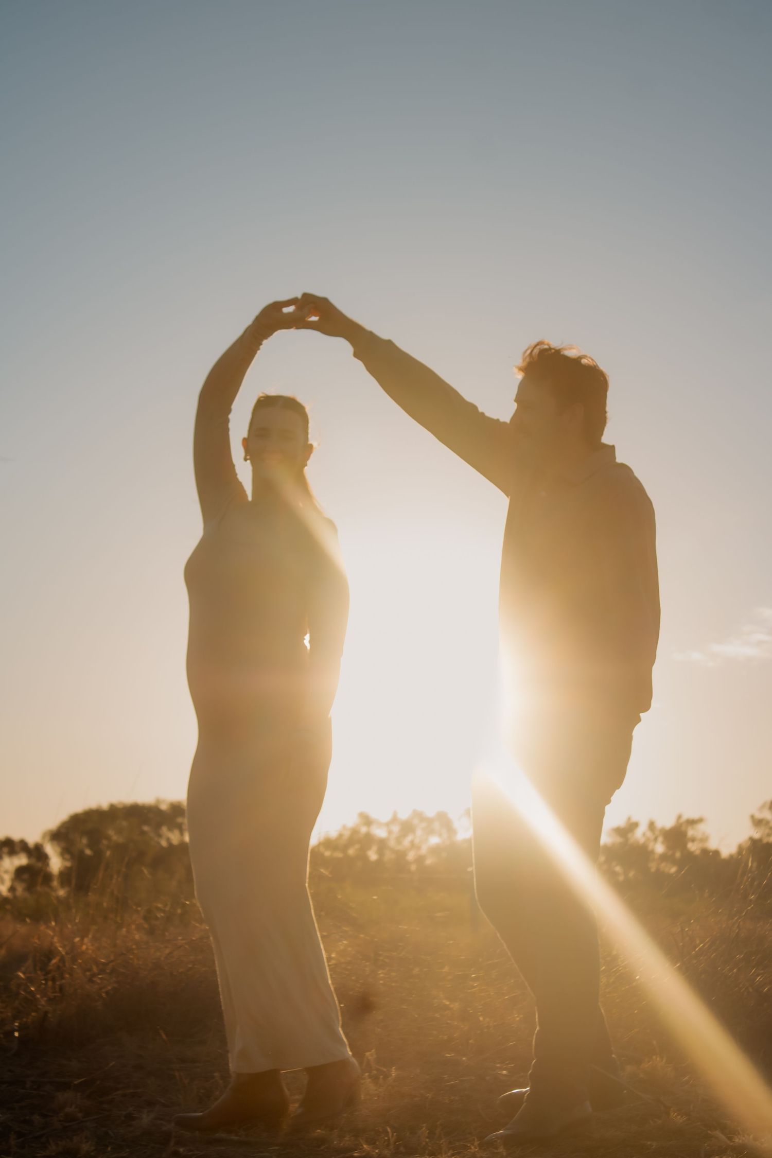Couple dancing silhouetted against golden sunset light, forming heart shape with raised hands outdoors.