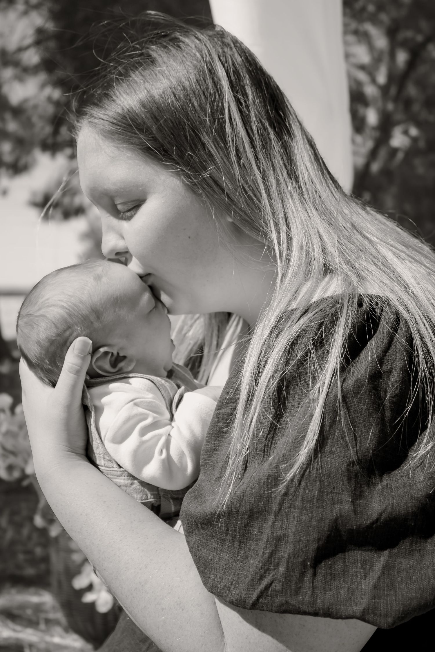 Black and white photo of a young mother tenderly kissing her newborn baby outdoors.