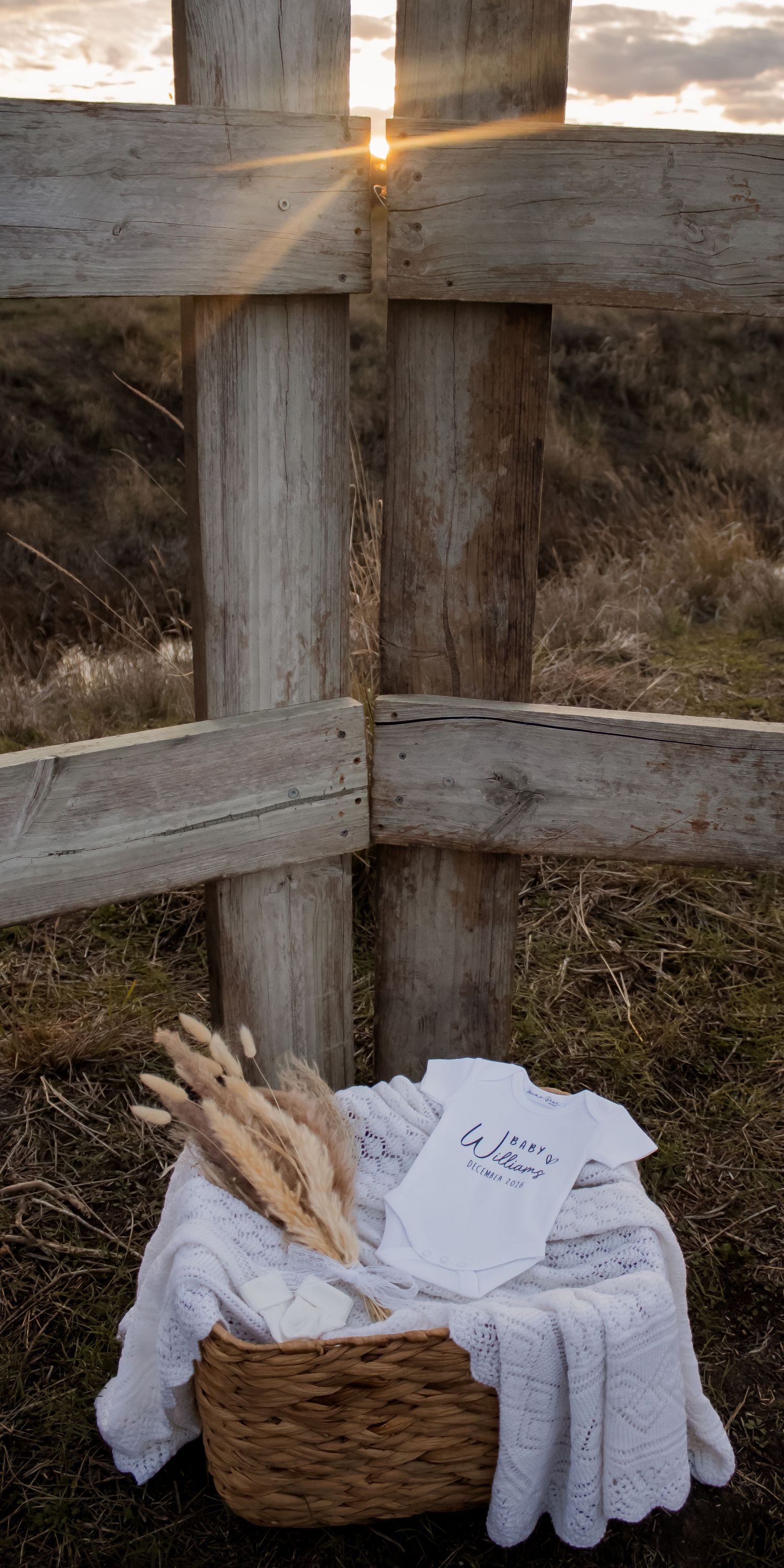 Rustic wooden cross at sunset with a wicker basket holding a white baby onesie, lace blanket, and dried pampas grass.