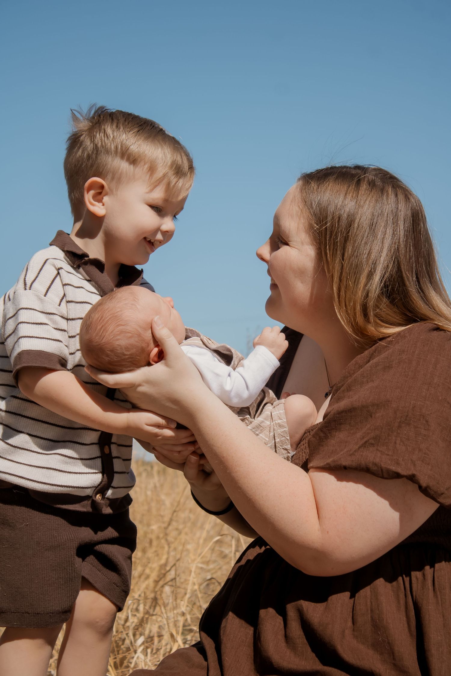 A mother and young boy joyfully hold a newborn baby outdoors in a sunny wheat field under a clear blue sky.