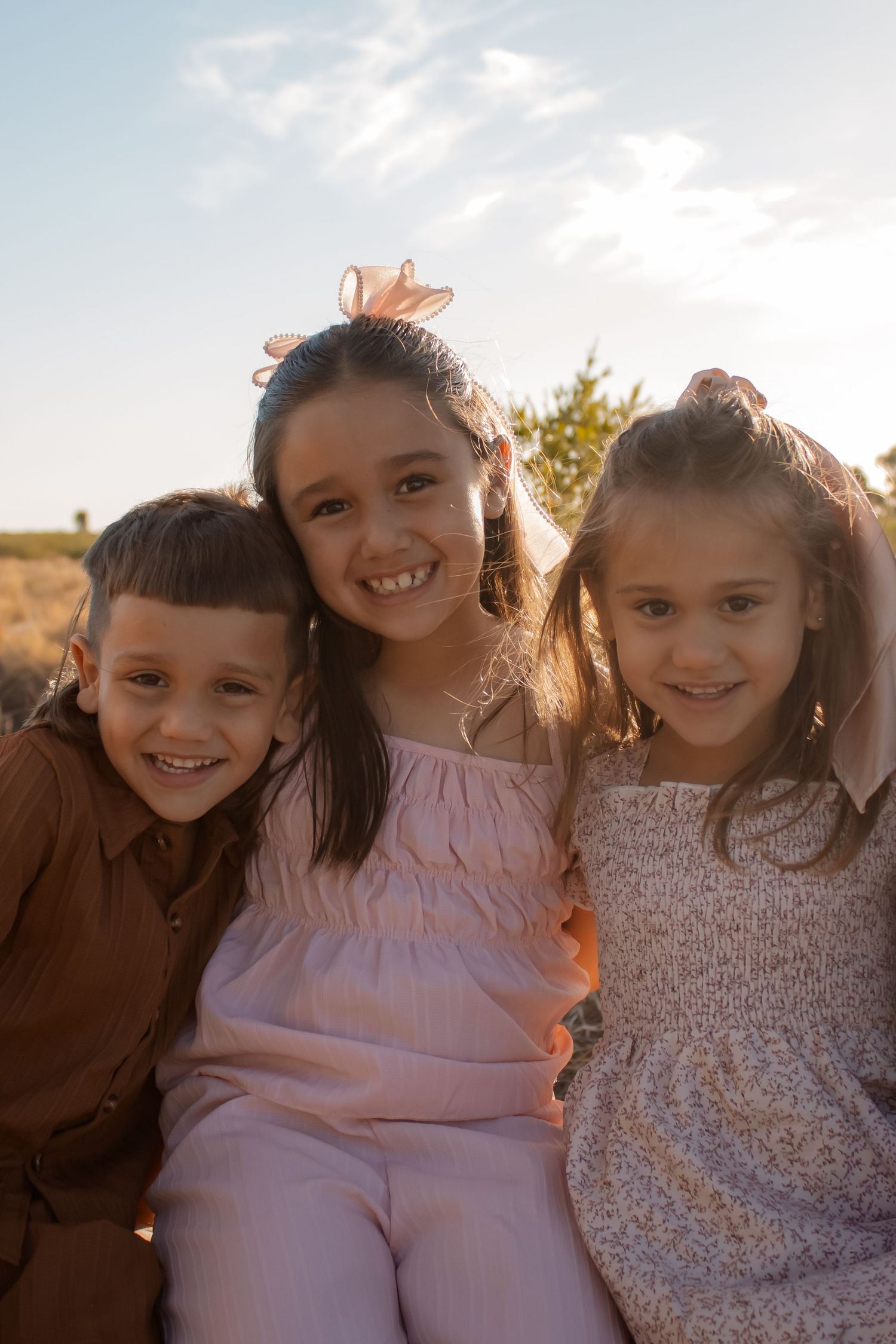 Three happy children smiling outdoors in golden sunlight, posing together in a sunny field.