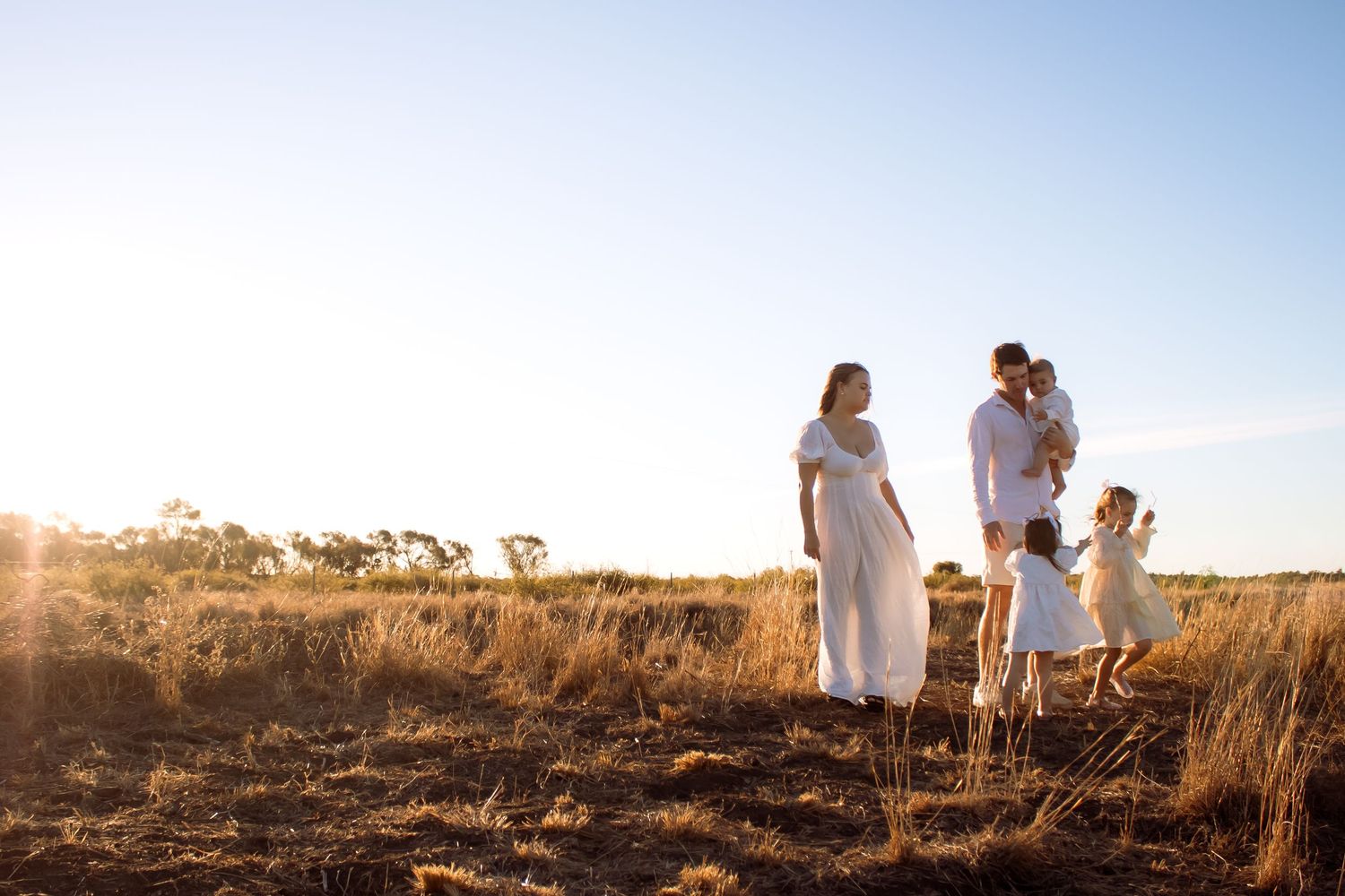 Family in white outfits standing in golden field at sunset, pregnant mother with partner holding child and toddler nearby.