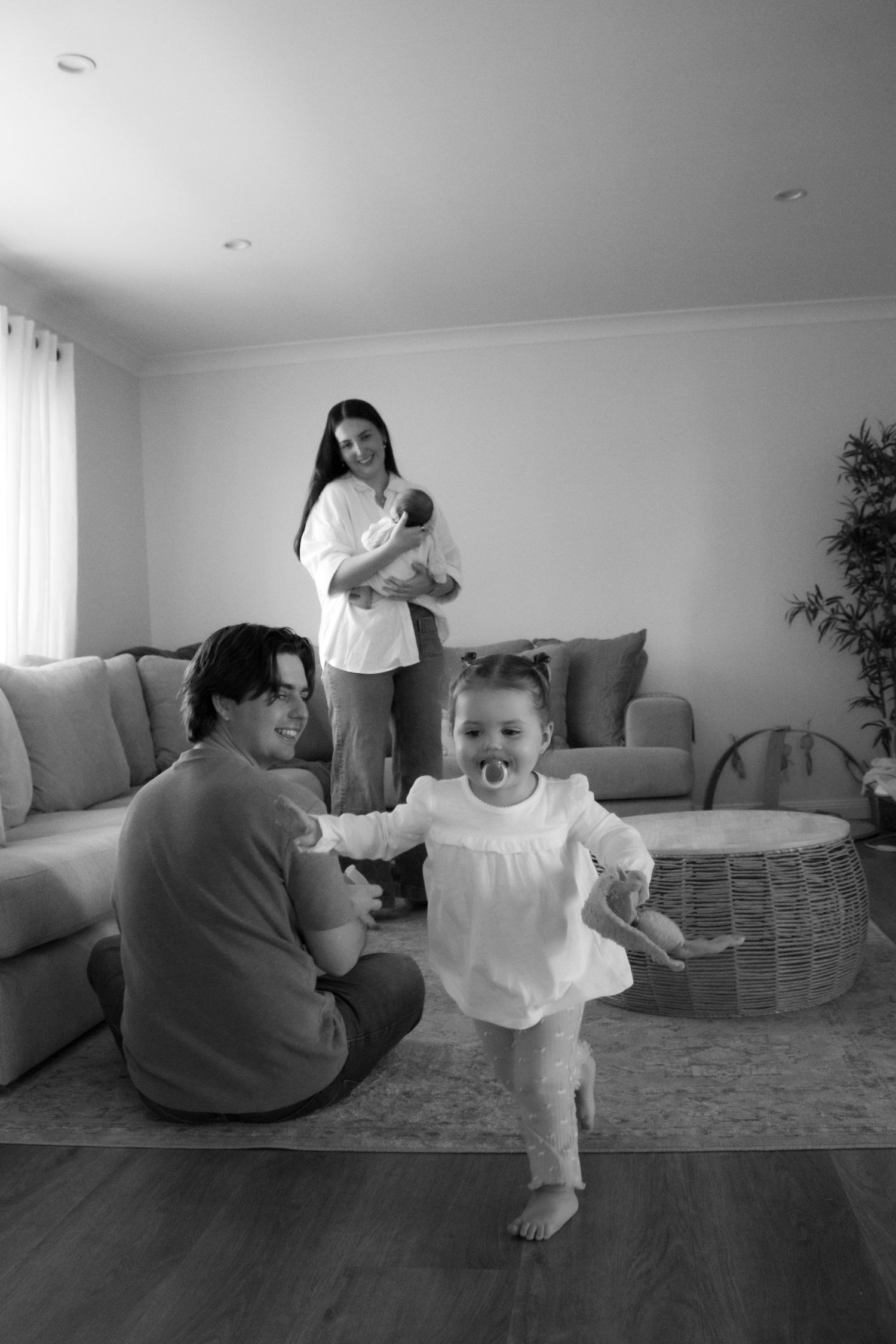 Black and white family photo with toddler walking, father sitting, and mother holding newborn in living room.